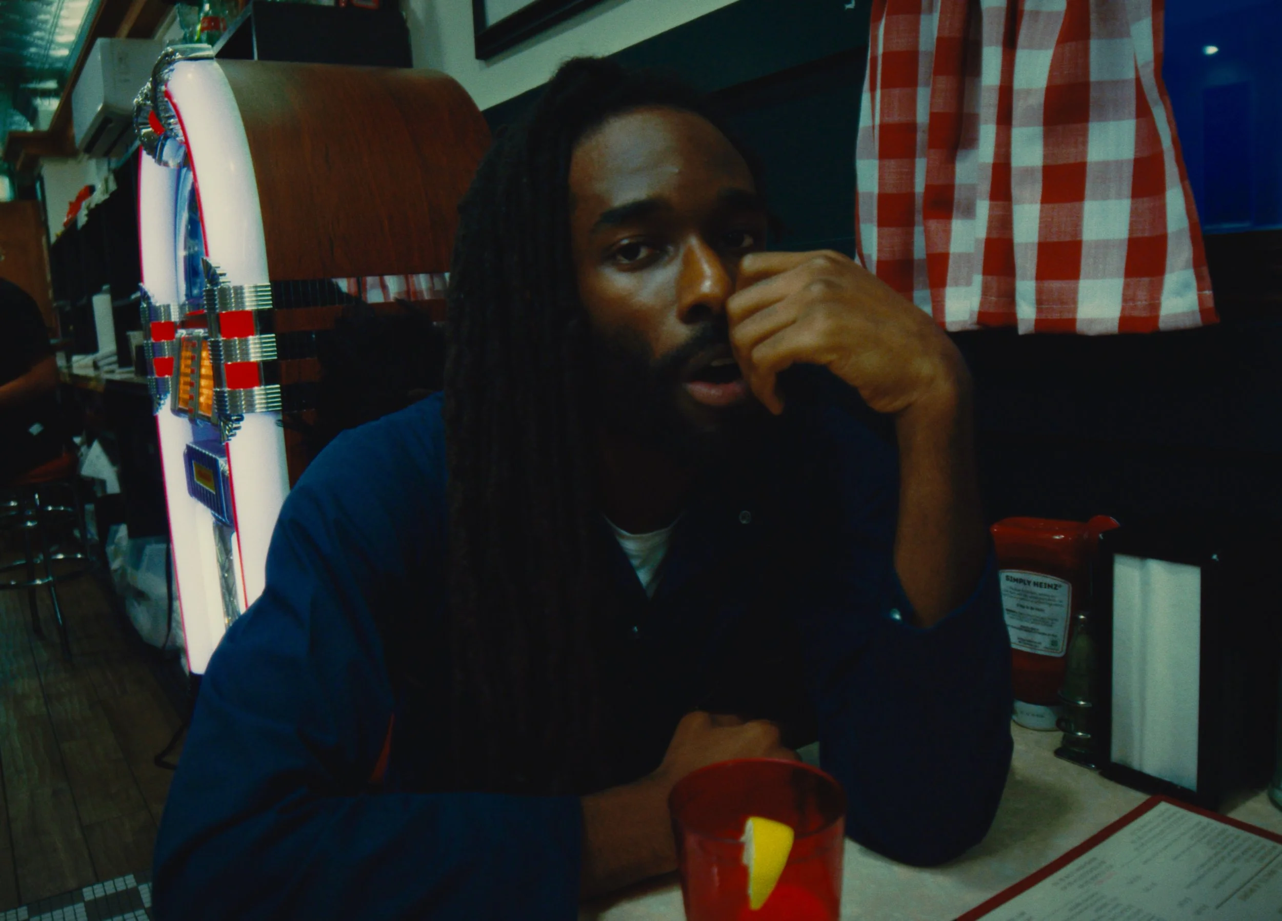 Man with long dreadlocks sitting at a restaurant table, resting his face on his hand, with a red cup in front of him and a vintage arcade game machine behind him.