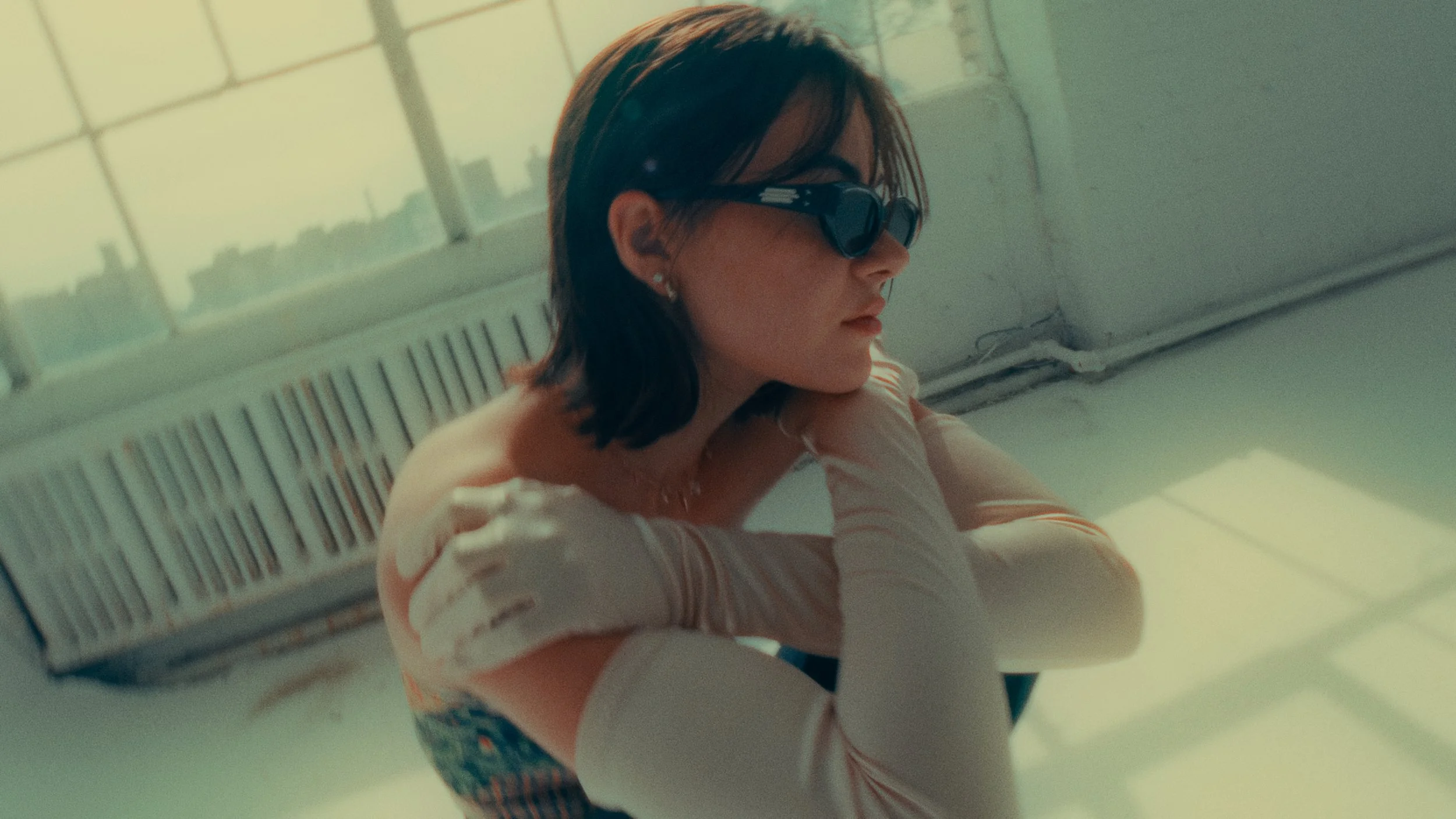 A woman with short dark hair wearing black sunglasses and gold hoop earrings, sitting by a window with city buildings in the background, illuminated by sunlight.