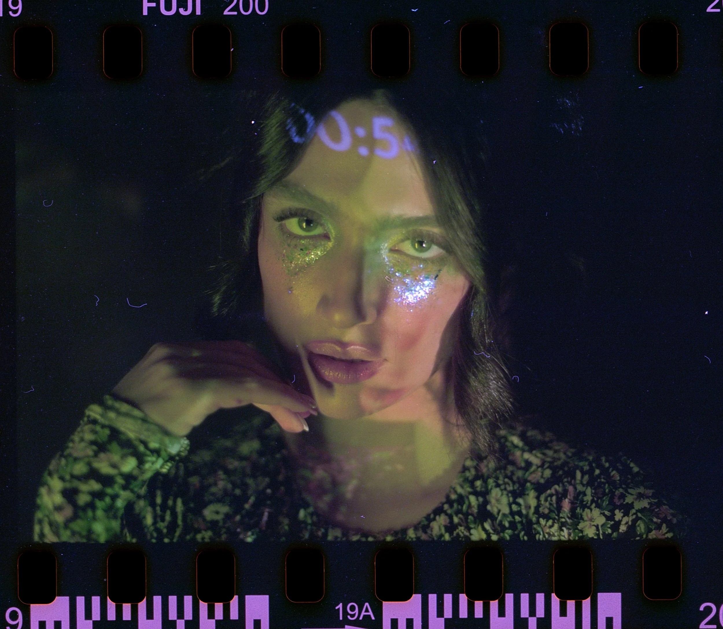 A woman with green eyes and glitter makeup on her face, posed with her hand near her chin, wearing a patterned shirt, illuminated by blacklight.editorial photoshoot for a brand