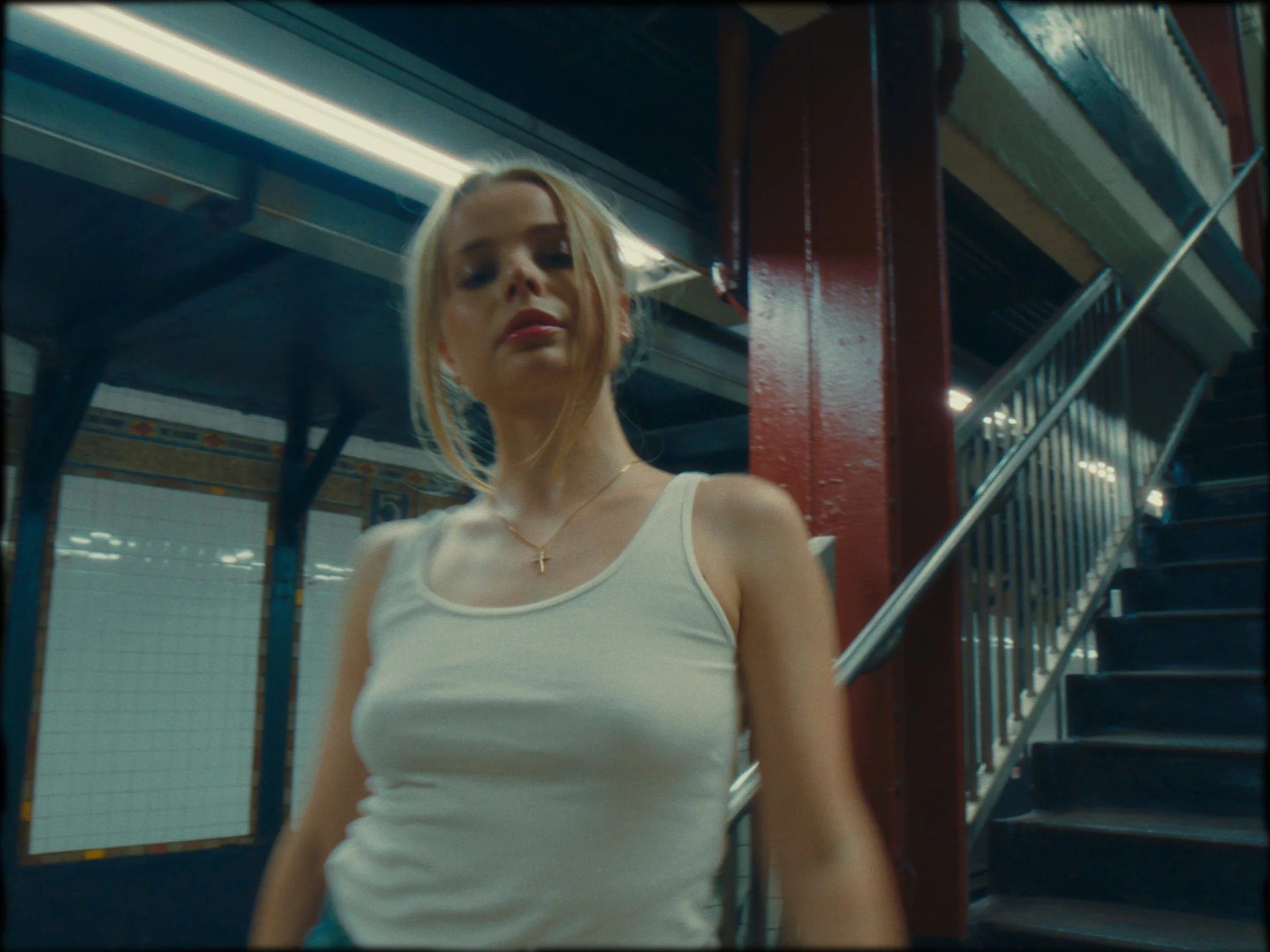 A young woman with blonde hair wearing a white tank top and a gold necklace standing on a subway platform.
