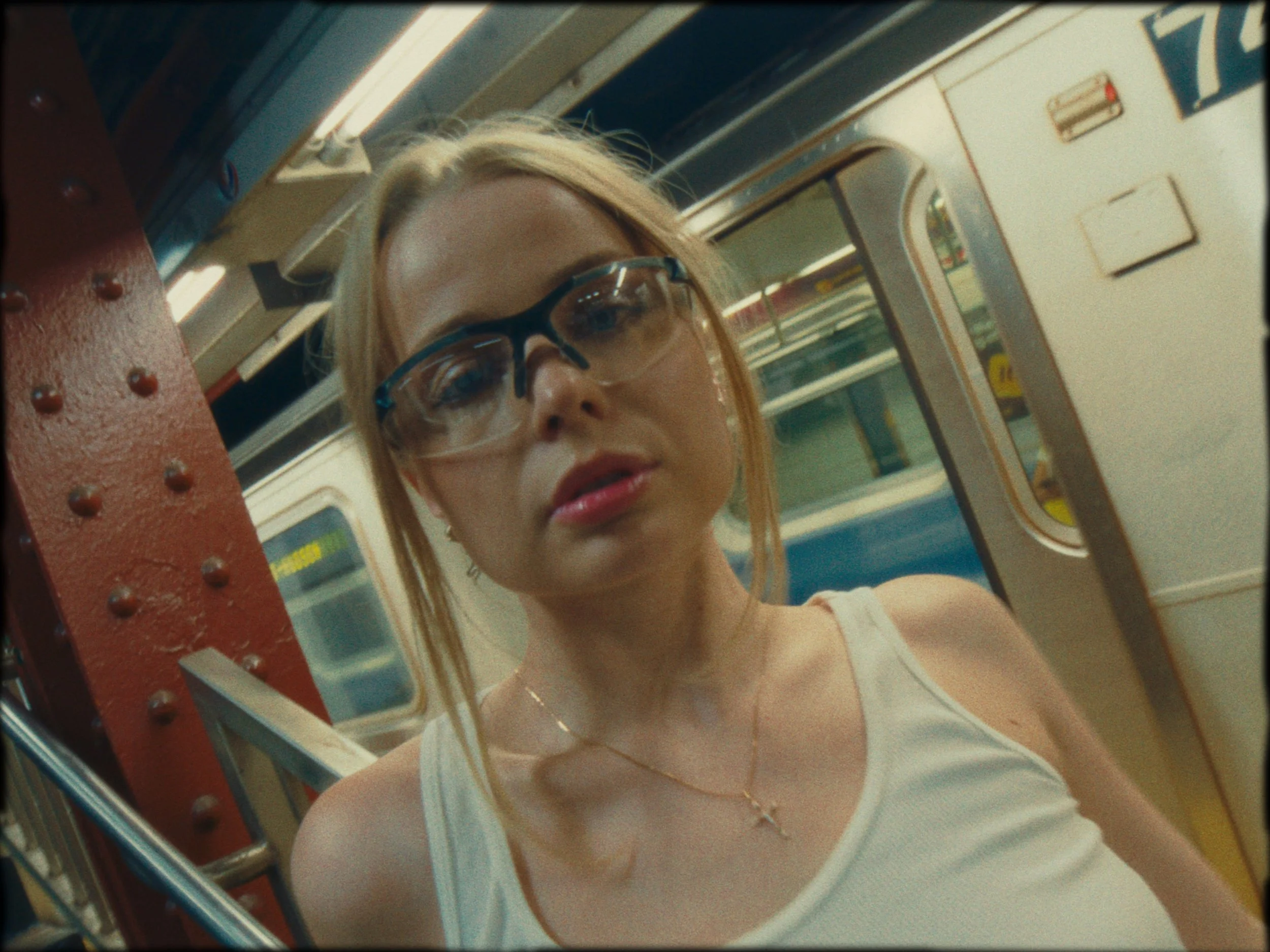 Close-up of a woman with blonde hair and glasses looking at the camera inside a subway train, with a metal railing and train door in the background.