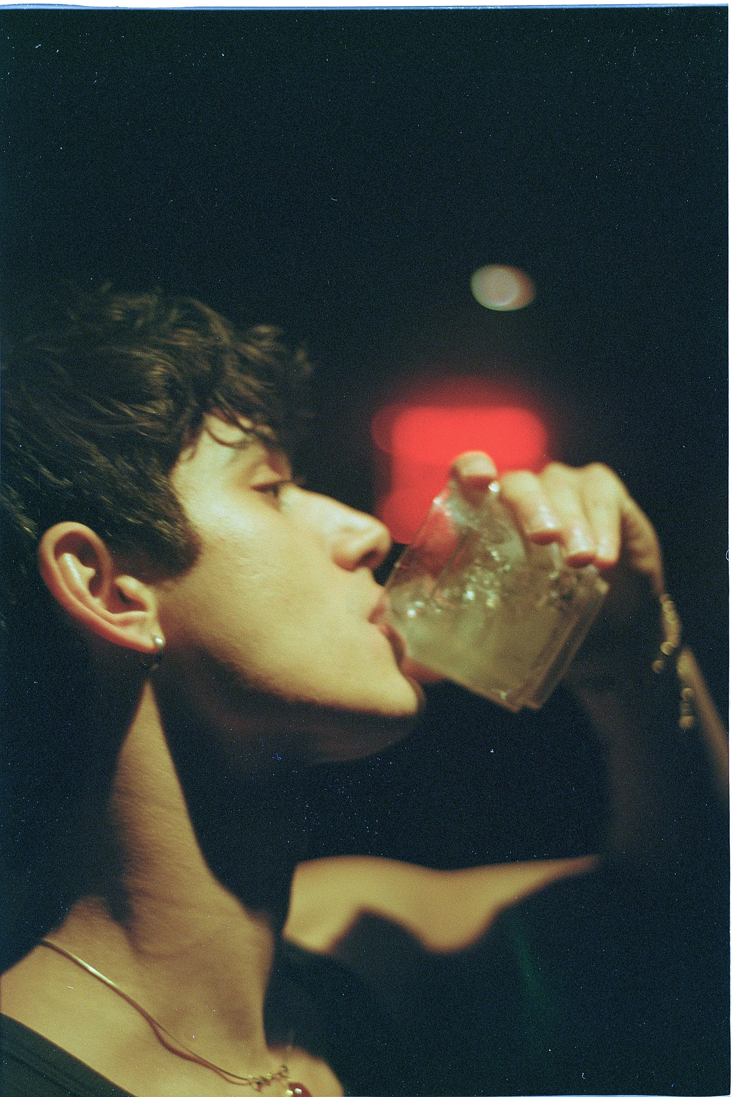 A young person with short, curly dark hair drinking from a glass of beverage in a dark setting with blurred red lights in the background.