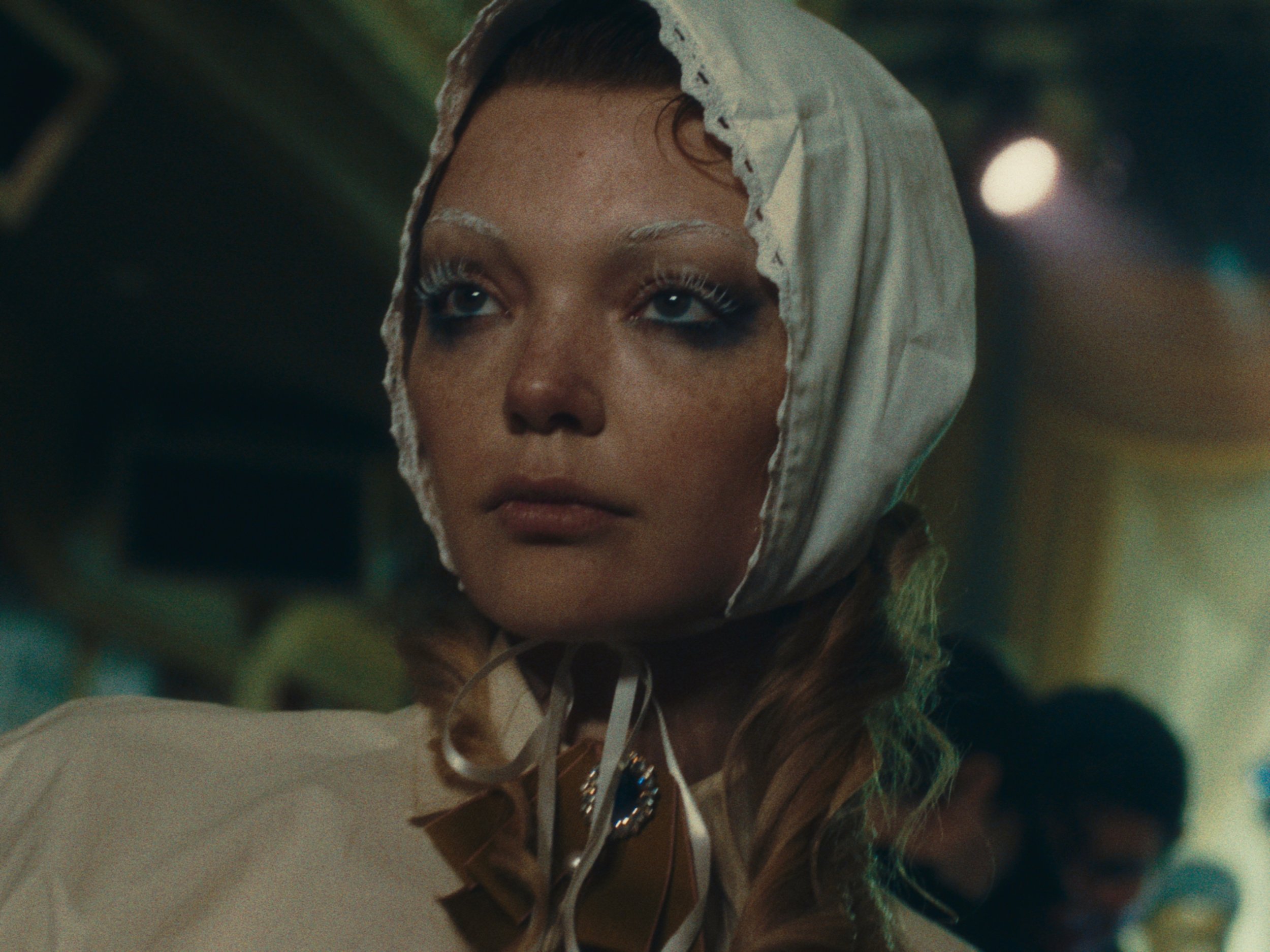 A woman with makeup and a cloth hood on her head looking slightly up and to the side, in a dimly lit environment.