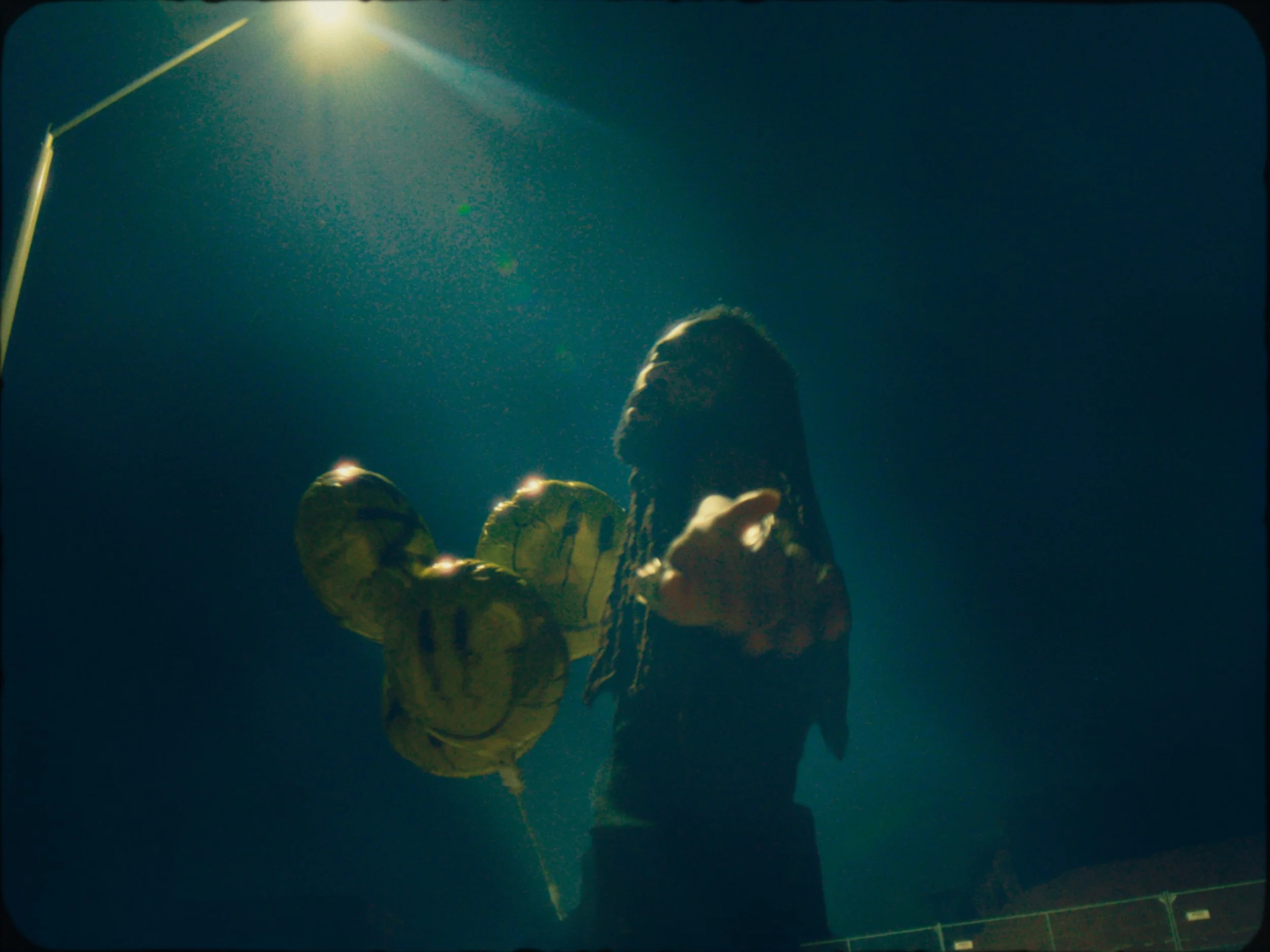 Person with dreadlocks holding a smiley face balloon at night under a streetlight.