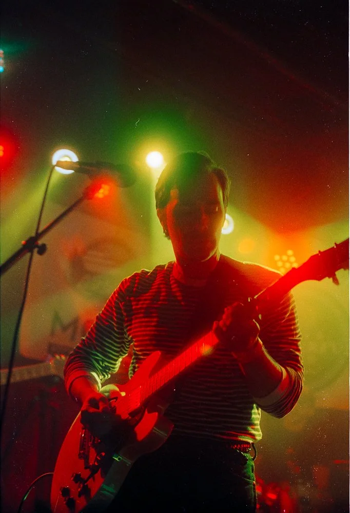 A musician playing an electric guitar on stage under colorful lights, with a silhouette of his face visible against the bright illumination.