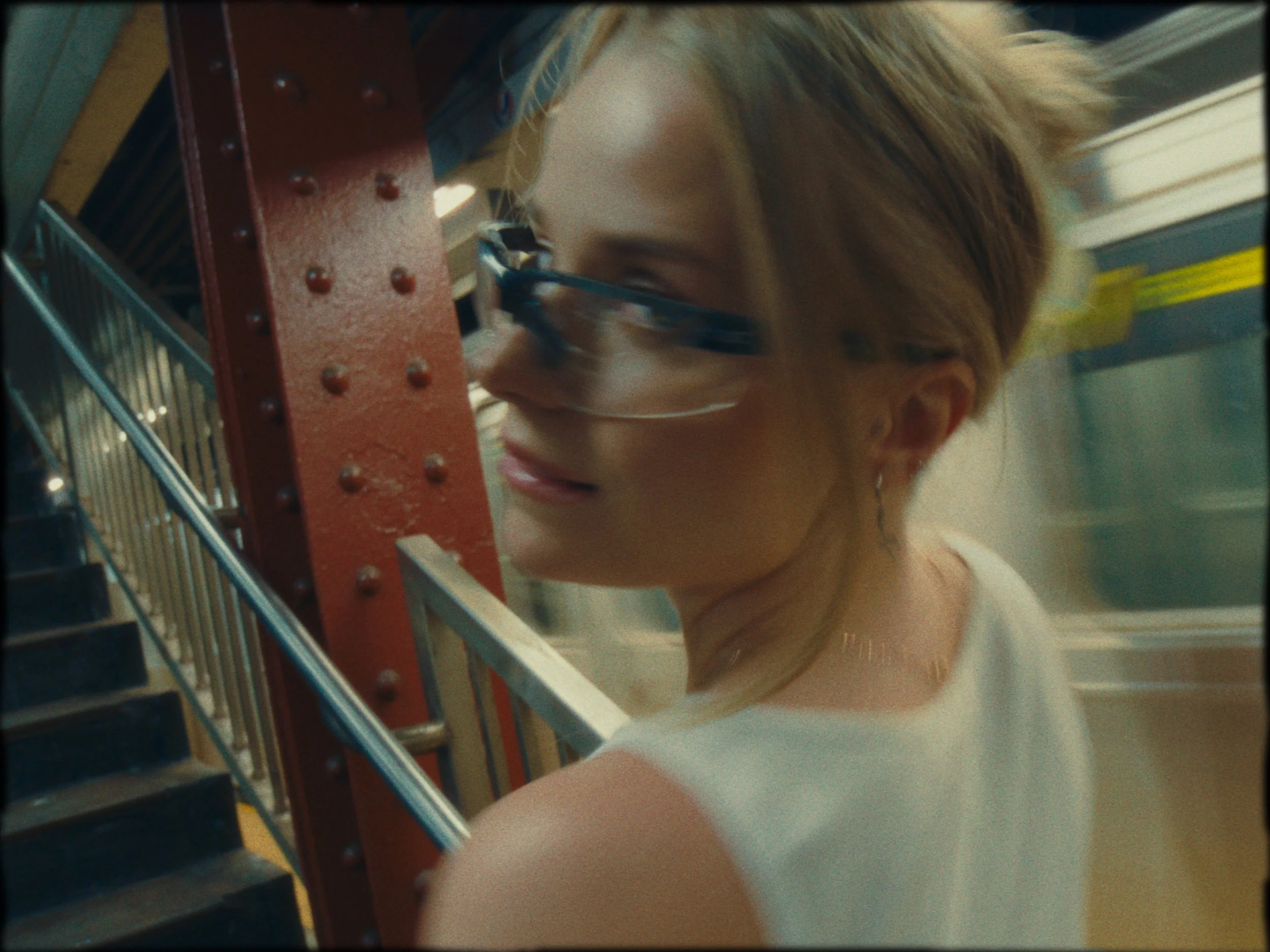 A woman with blonde hair and sunglasses, wearing a white top, leaning against a metal railing on a staircase in an urban environment.