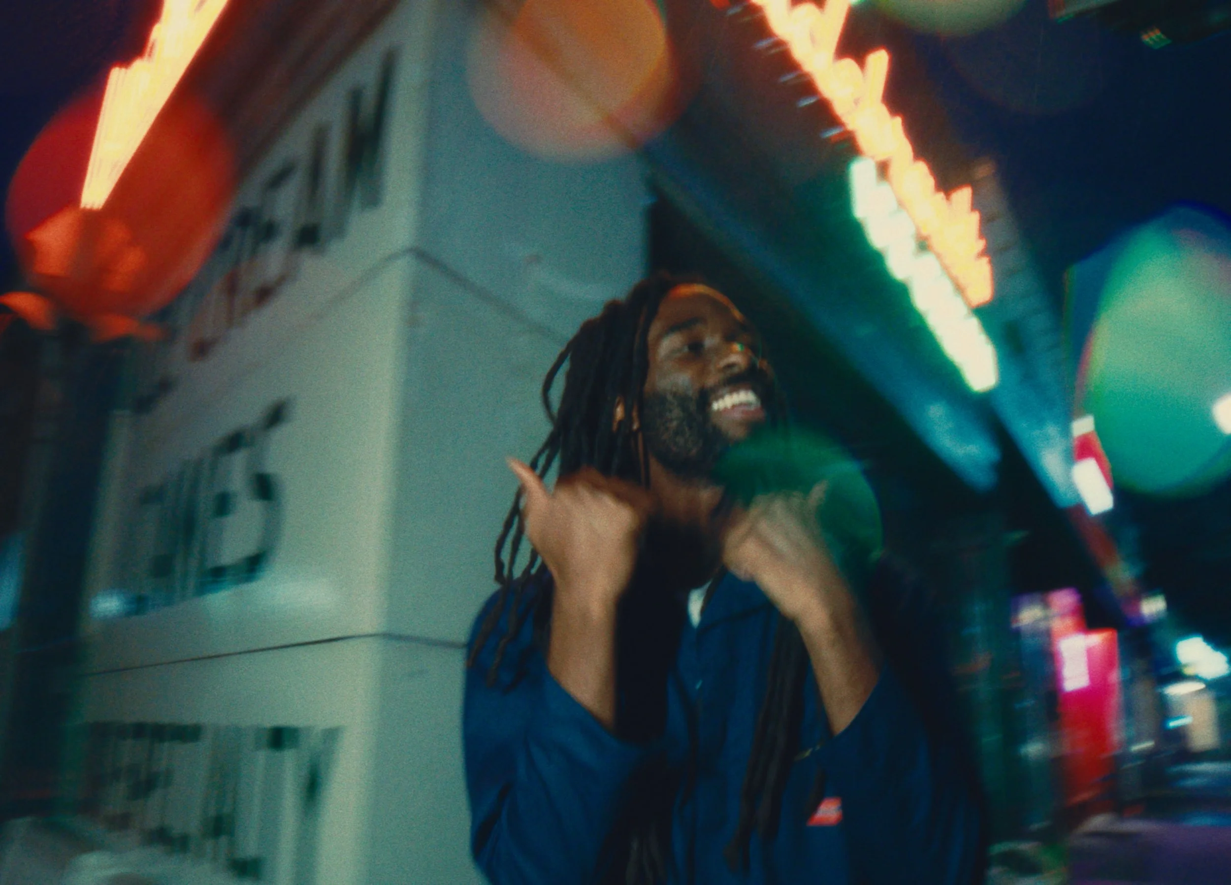 A man with dreadlocks and a beard smiling and touching his necks under neon lights at night, with blurred city signs in the background.