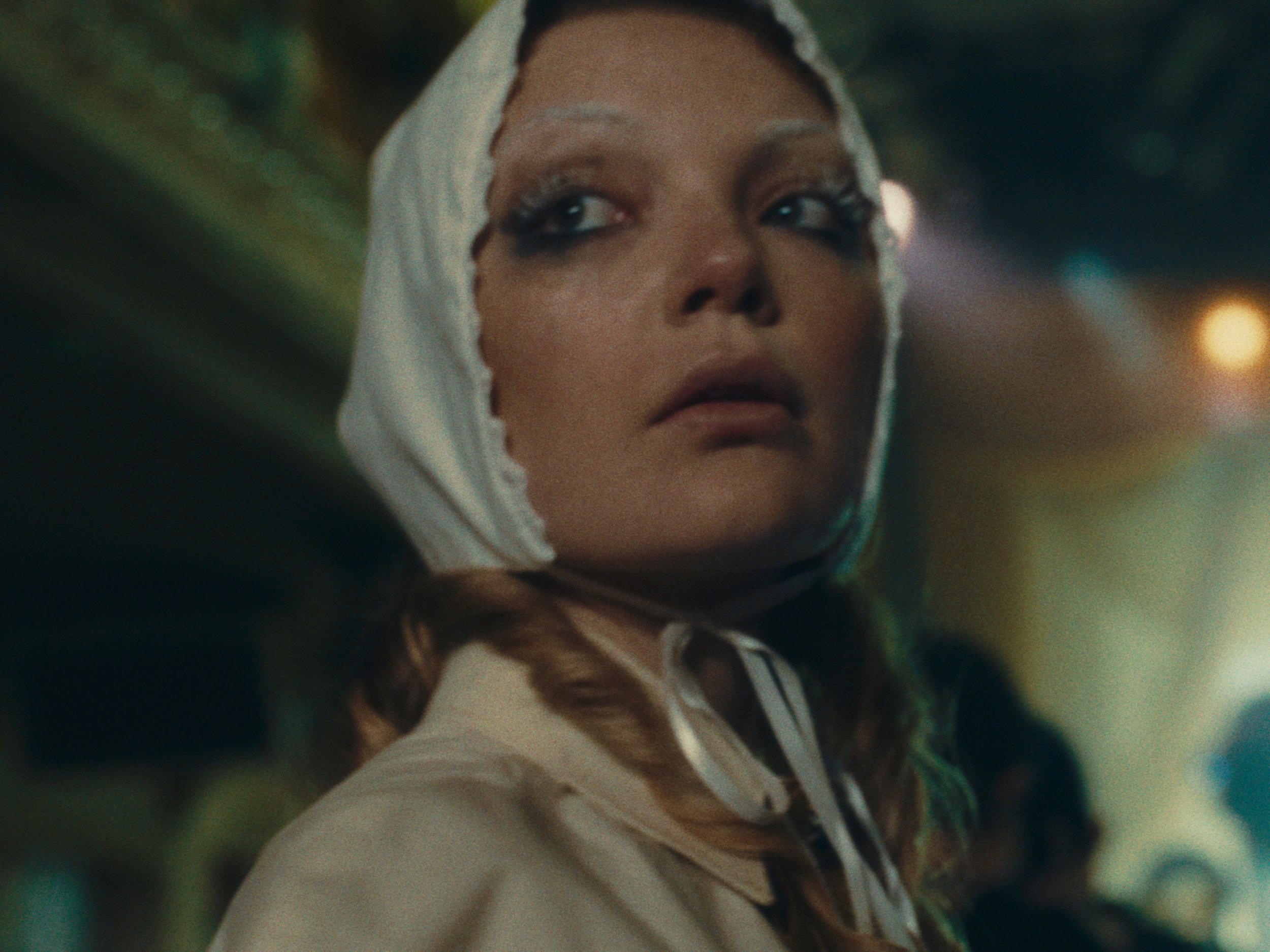 A woman with styled hair and makeup wearing a beige coat and a white headscarf in a dimly lit environment.