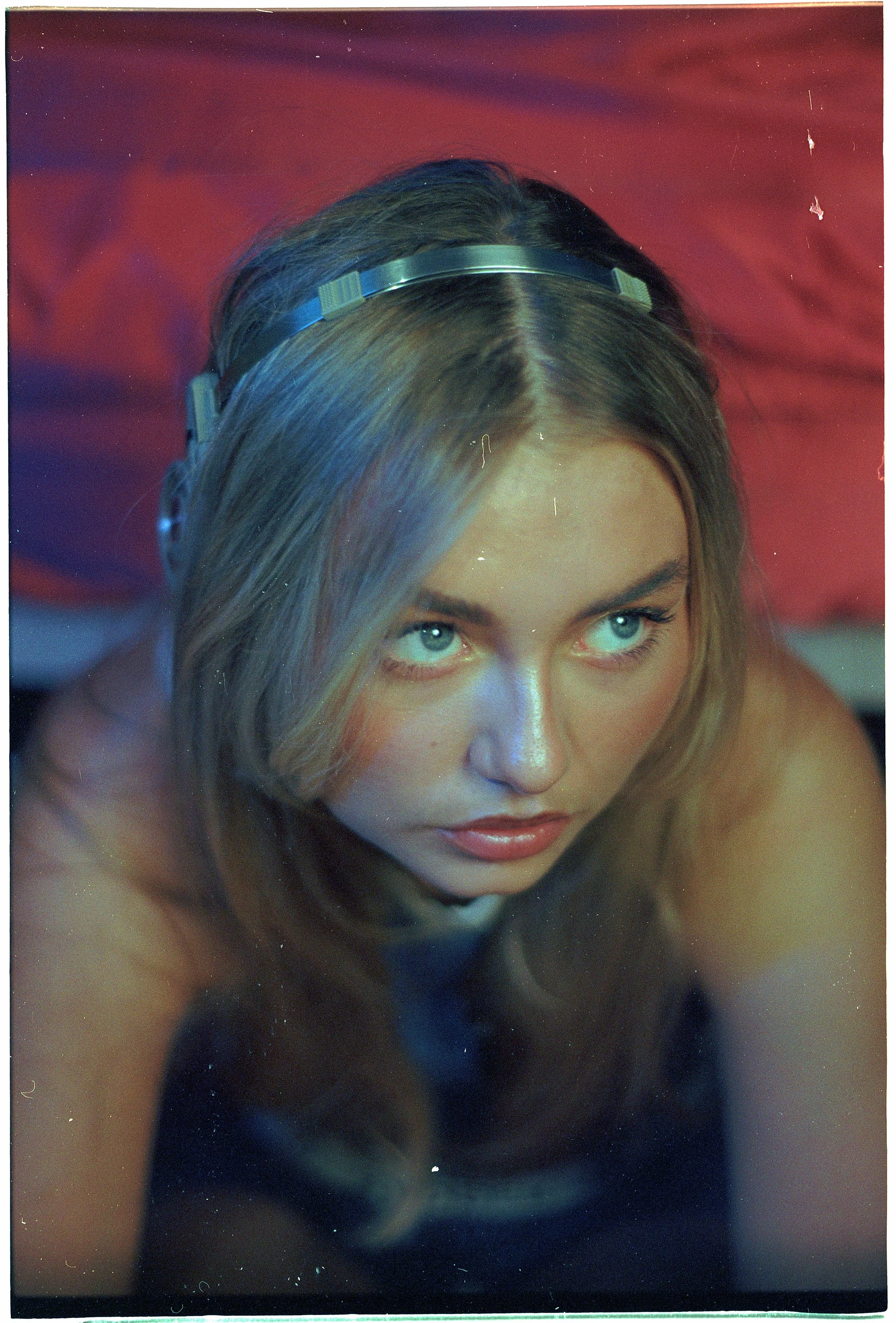 A woman with long, wavy hair wearing headphones looking upward with intense blue eyes, in front of a colorful, blurred background.