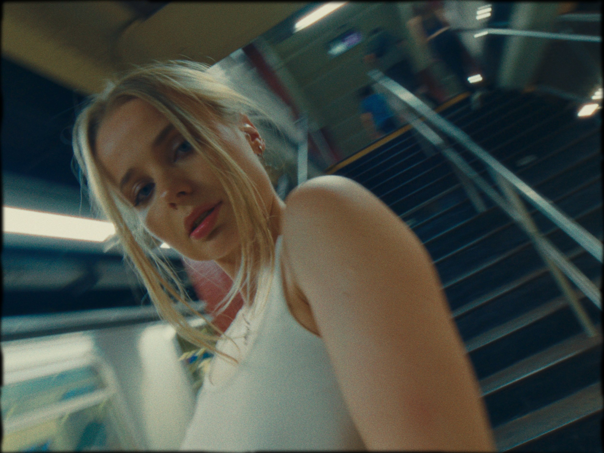 A young woman with blonde hair wearing a white sleeveless top is sitting on a bench in what appears to be a train or subway station, looking at the camera with a neutral expression.