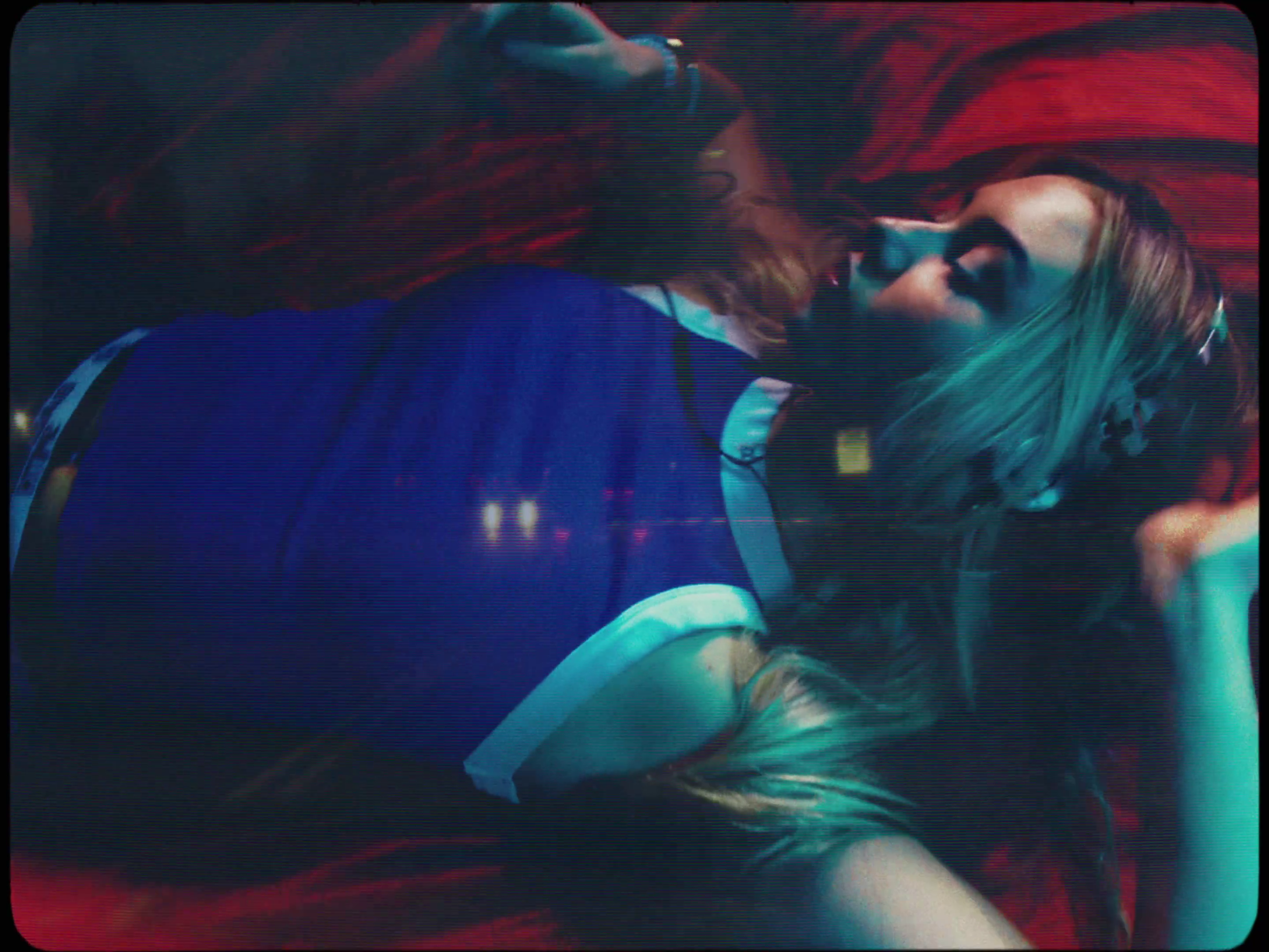 Two girls lying on a red bed, one with colorful hair and the other with dark hair, facing each other closely.