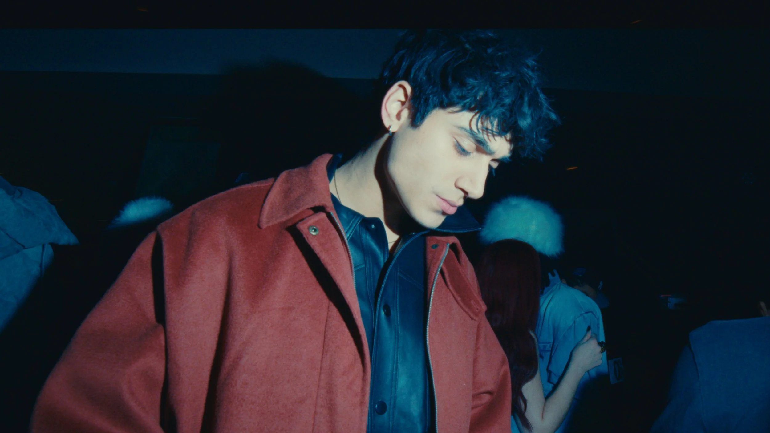 A young man with curly dark hair is looking down with a calm expression, wearing a dark red coat over a black leather jacket, in a dimly lit setting with other people in the background.