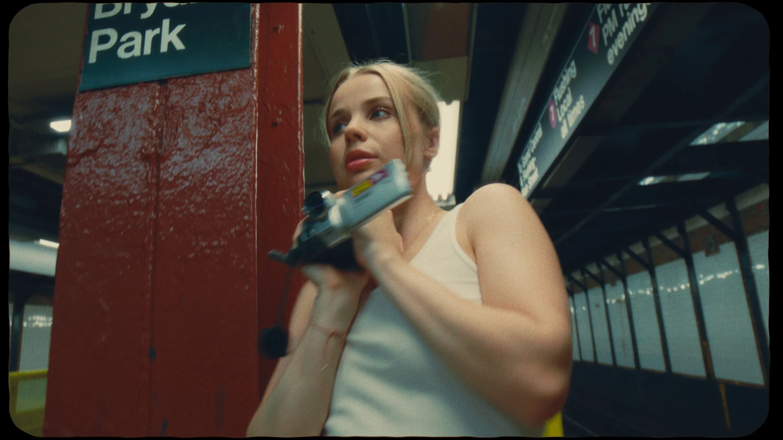 A young woman in a white tank top holding a gun in a subway station with a red pillar and dark ceiling panels.