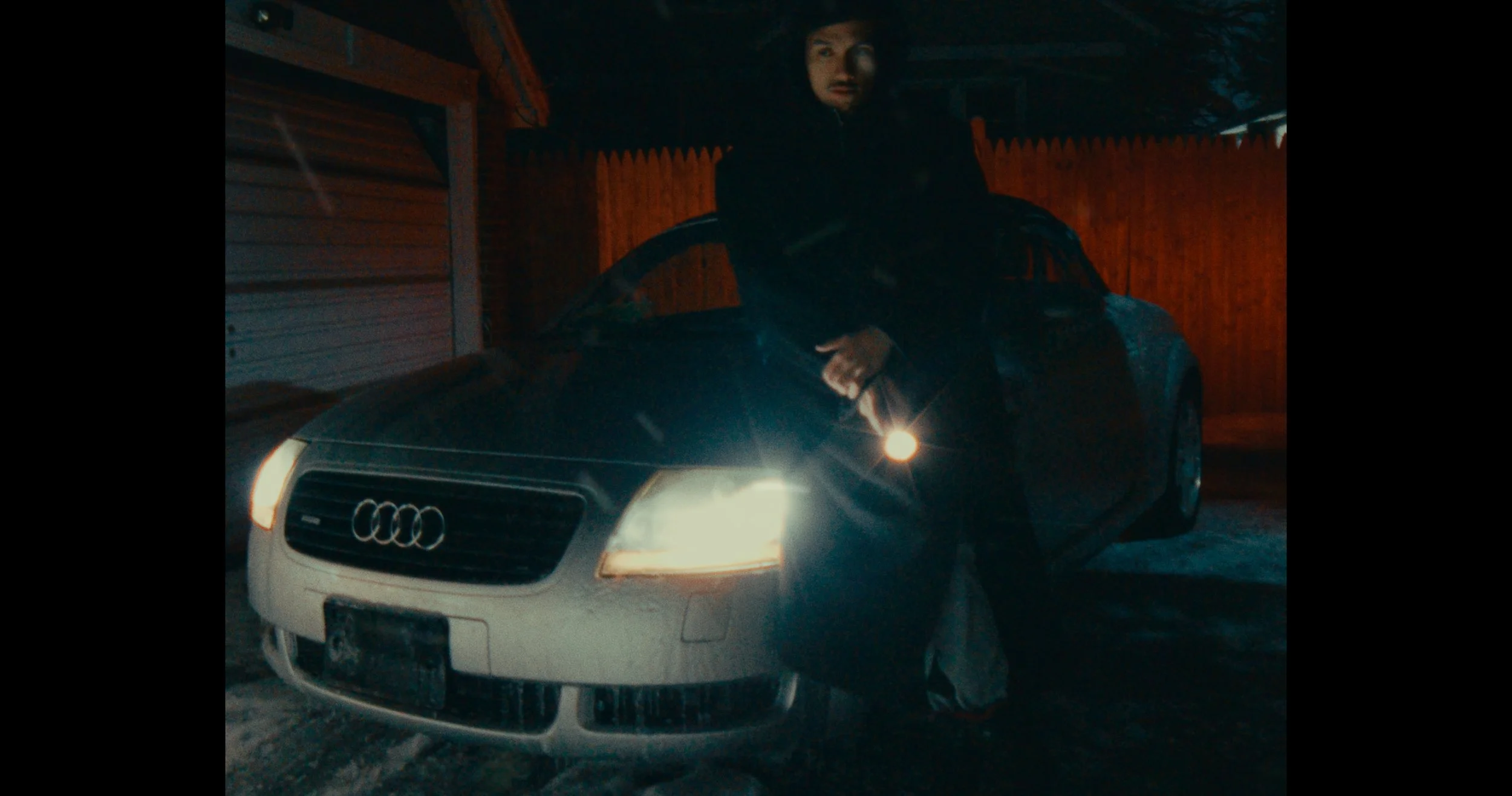 A person in dark clothing standing in front of a black Audi car, holding a flashlight, in a dimly lit outdoor area at night with a wooden fence in the background.