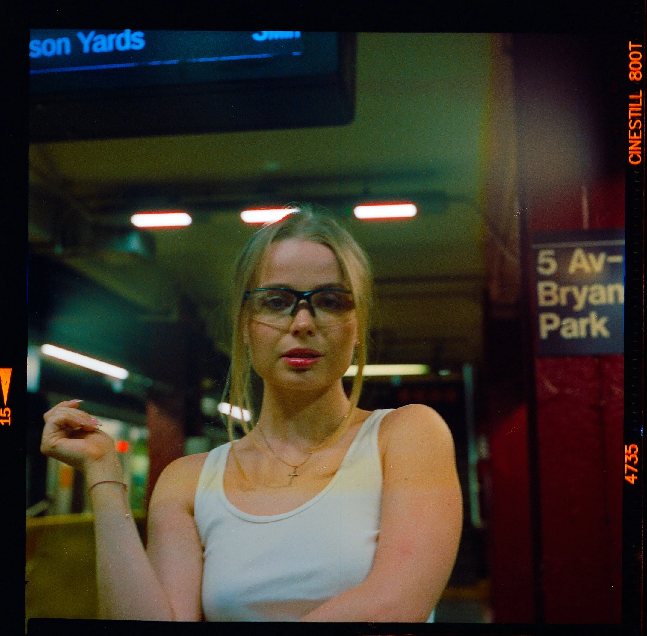 A woman with blonde hair, glasses, wearing a white tank top, standing in a subway station. She is looking at the camera with her right hand raised near her shoulder. There is an exit sign that reads '5 Av - Bryan Park' to her right. The background includes station lighting, a ceiling, and part of an electronic display.