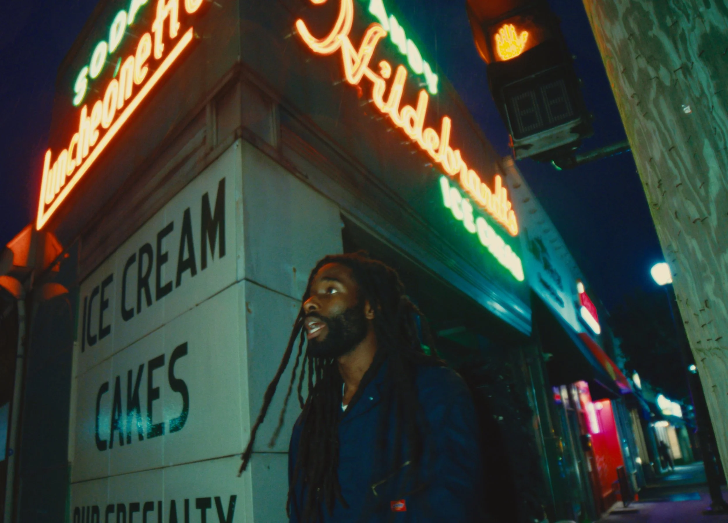 A man with dreadlocks and a beard standing on a city street at night, illuminated by colorful neon signs advertising ice cream, cakes, and specialty drinks.