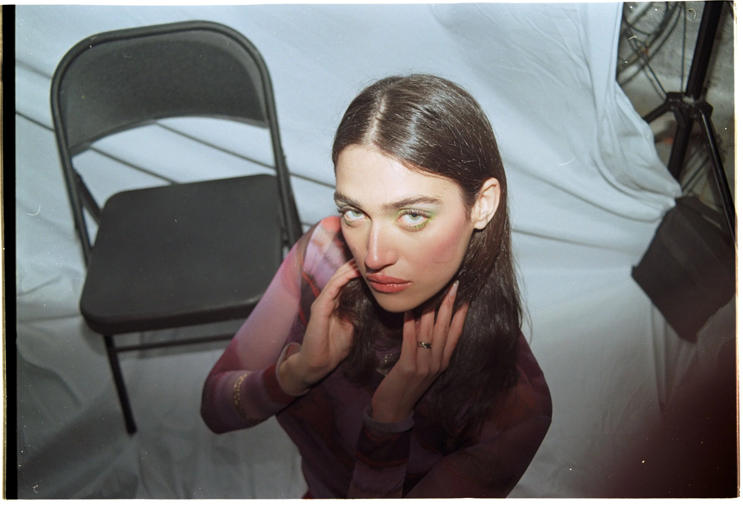 A woman with long dark hair and green eyeshadow sitting at a table, looking up at the camera, with a black folding chair and a black bag in the background.editorial photoshoot for a brand