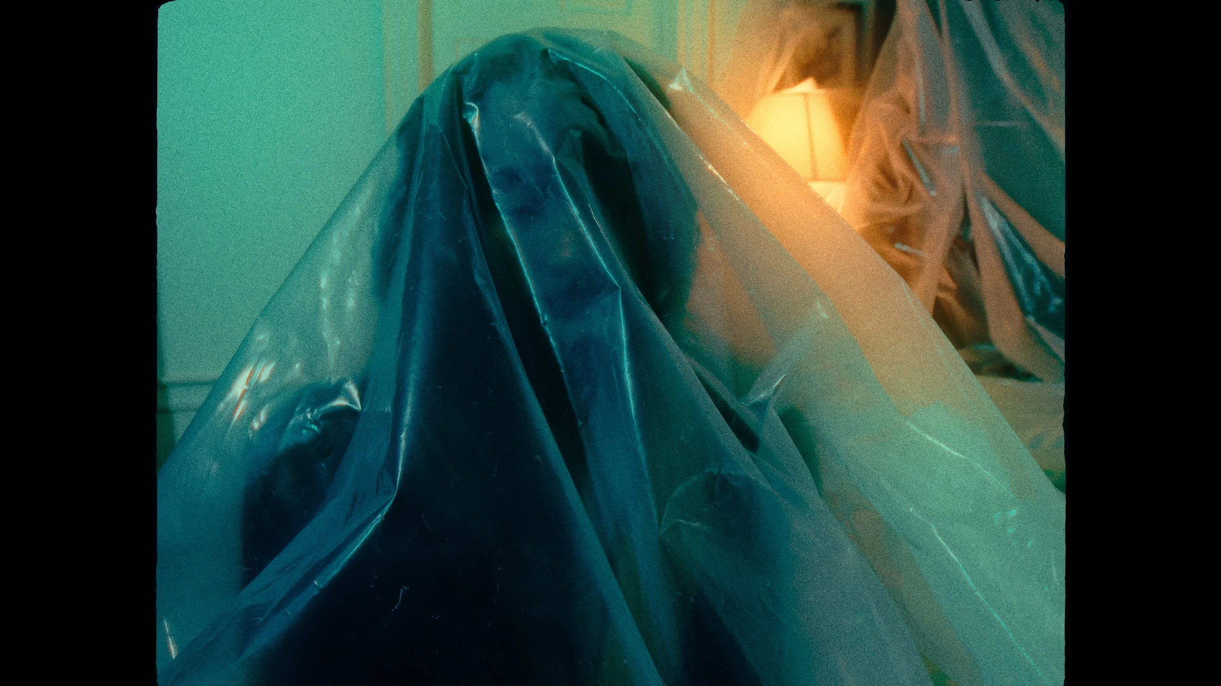 A person covered with a large, transparent plastic sheet in a dimly lit room with a lamp in the background.