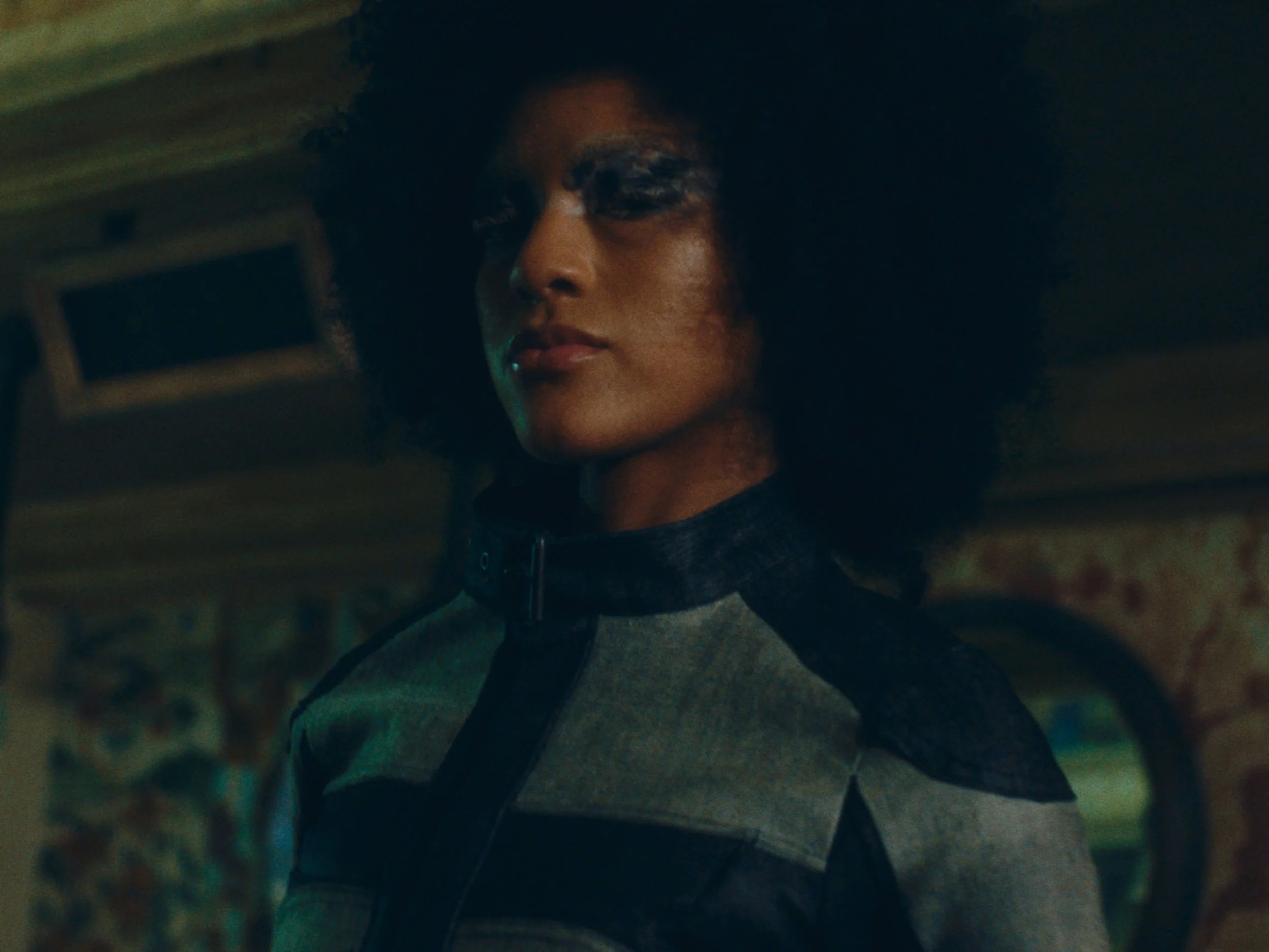 Close-up of a woman with dark curly hair, wearing a black and grey tactical suit with a high collar, standing in a dimly lit environment.