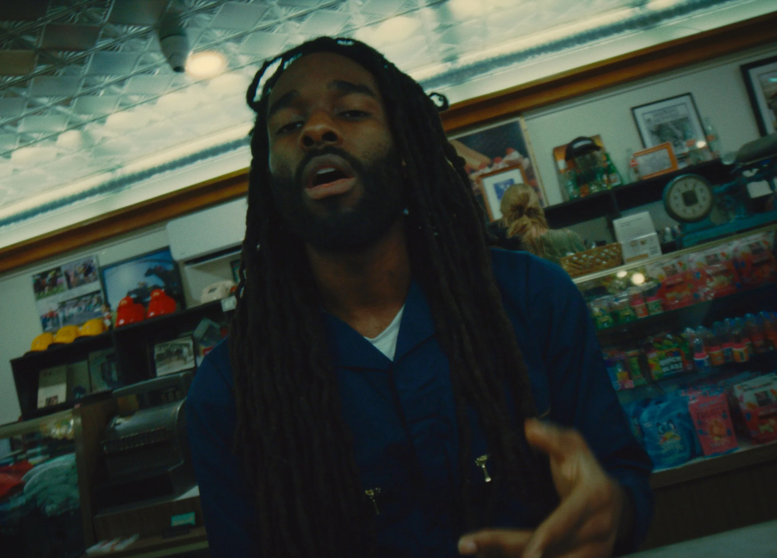 A man with long dreadlocks and a beard is in a store, facing the camera with a slightly open mouth. Behind him are shelves with various products, including snacks and beverages, and a woman with blond hair sits at the counter.