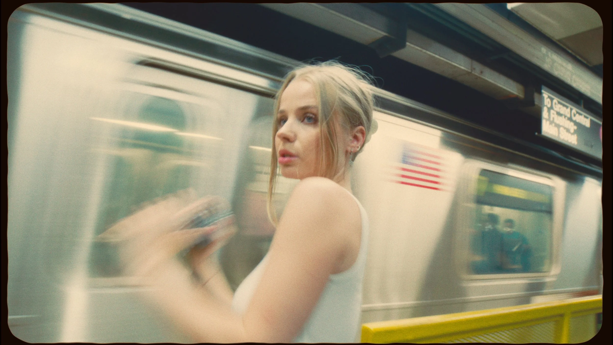 Young woman with blonde hair in a bun wearing a white tank top, standing on a subway platform, with a moving train in the background.