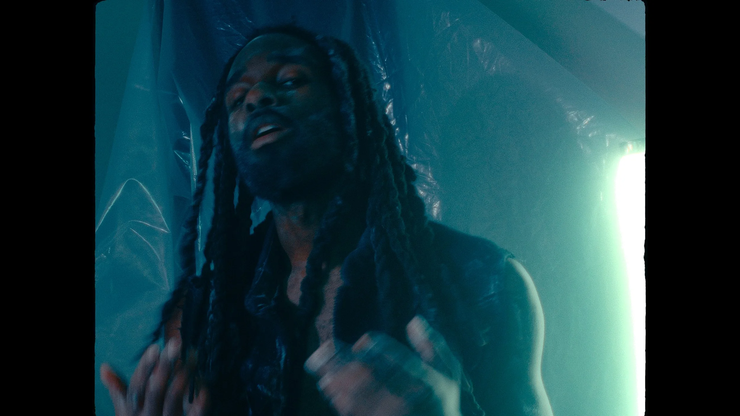 A man with long braided hair and a beard, wearing a sleeveless leather vest, standing in front of a bright light source with a blue and green hue, with reflective metallic surfaces in the background.