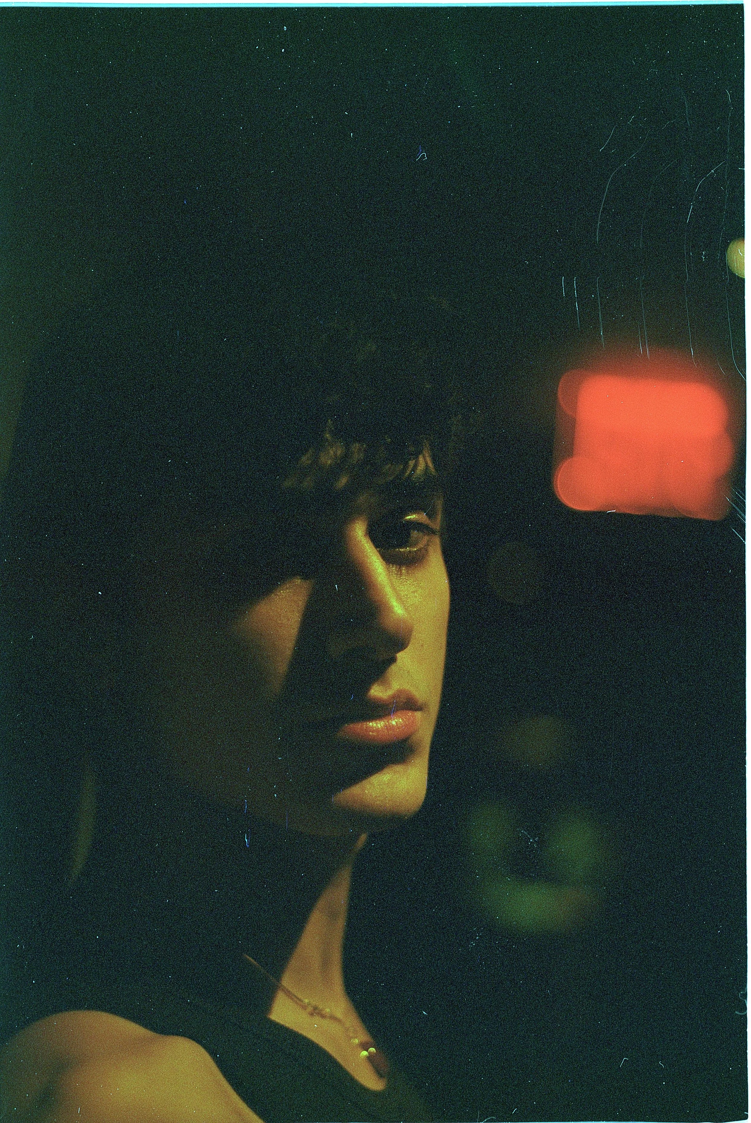 A woman's face illuminated by green and yellow lighting, with a blurred red and orange background, and a dark environment.