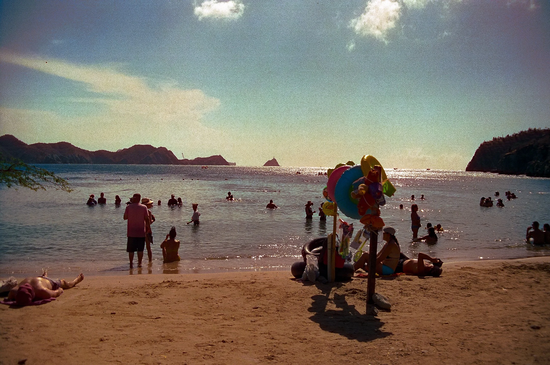 People enjoying a beach with the ocean and distant islands in the background, colorful beach toys and an inflatable ring on the sand in the foreground, some people wading in the water, and others relaxing on the shore.