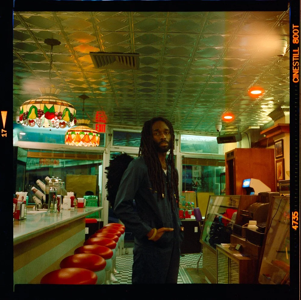 A man standing inside a retro-style diner with red barstools, colorful hanging lamps, and a checkered floor, facing the camera with his hands in his pockets.
