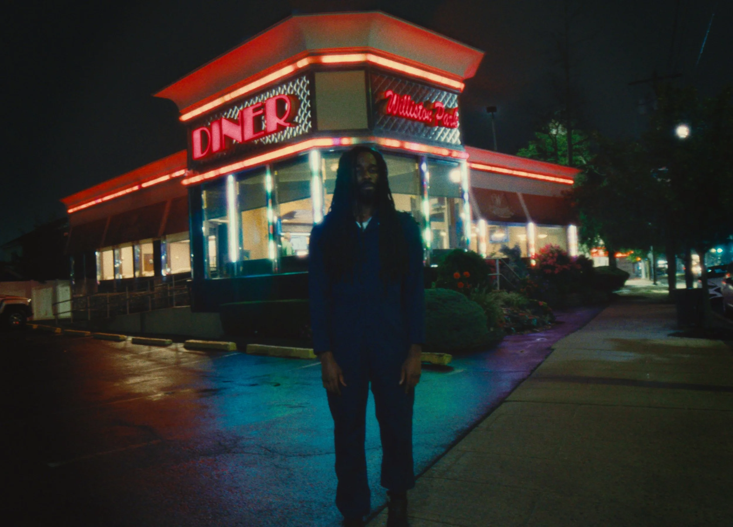 Person with long dreadlocks standing outside a neon-lit diner at night.