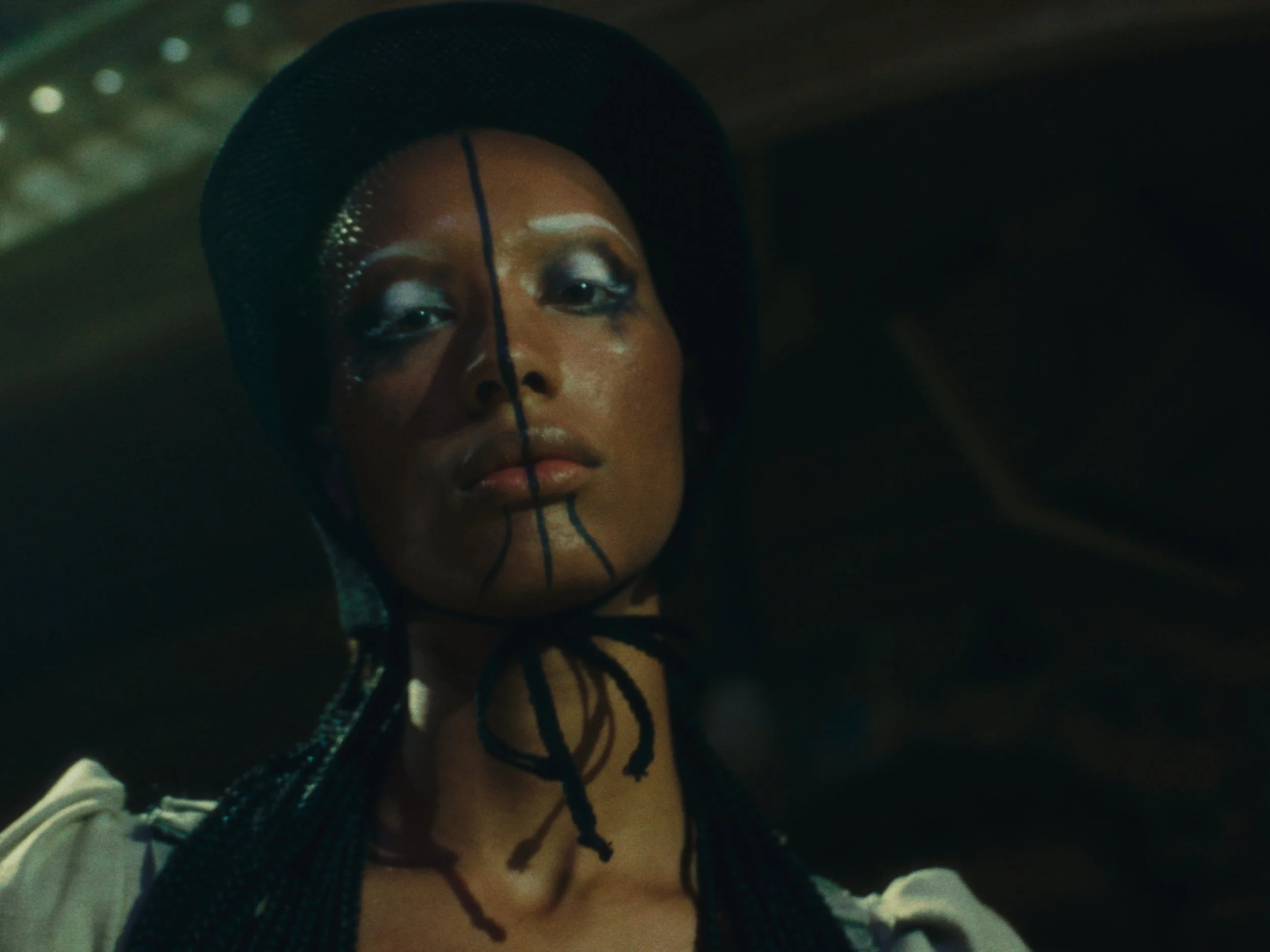 Close-up of a woman with elaborate face makeup, wearing a black hat with a netted veil, and braided hair, looking down.
