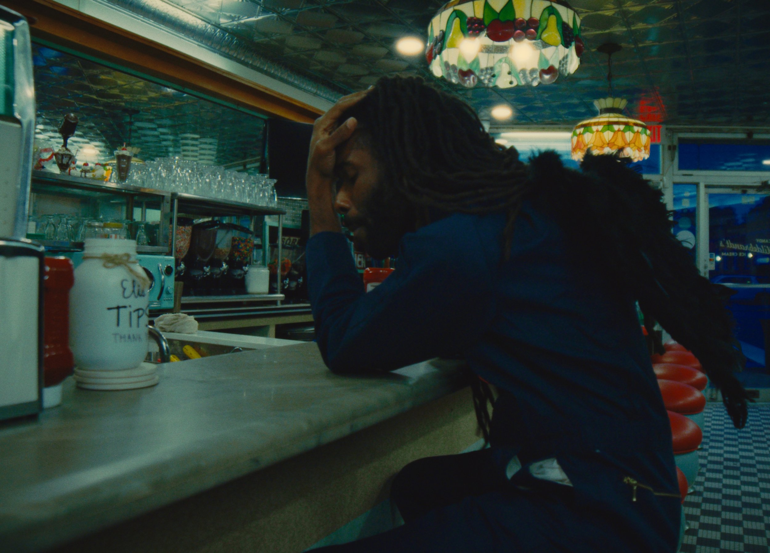 A man with long dreadlocks sitting at a diner counter, resting his head in his hand in a moment of frustration or exhaustion. The diner has retro lighting and decorated lamps, with various glassware and condiments visible on the counter.