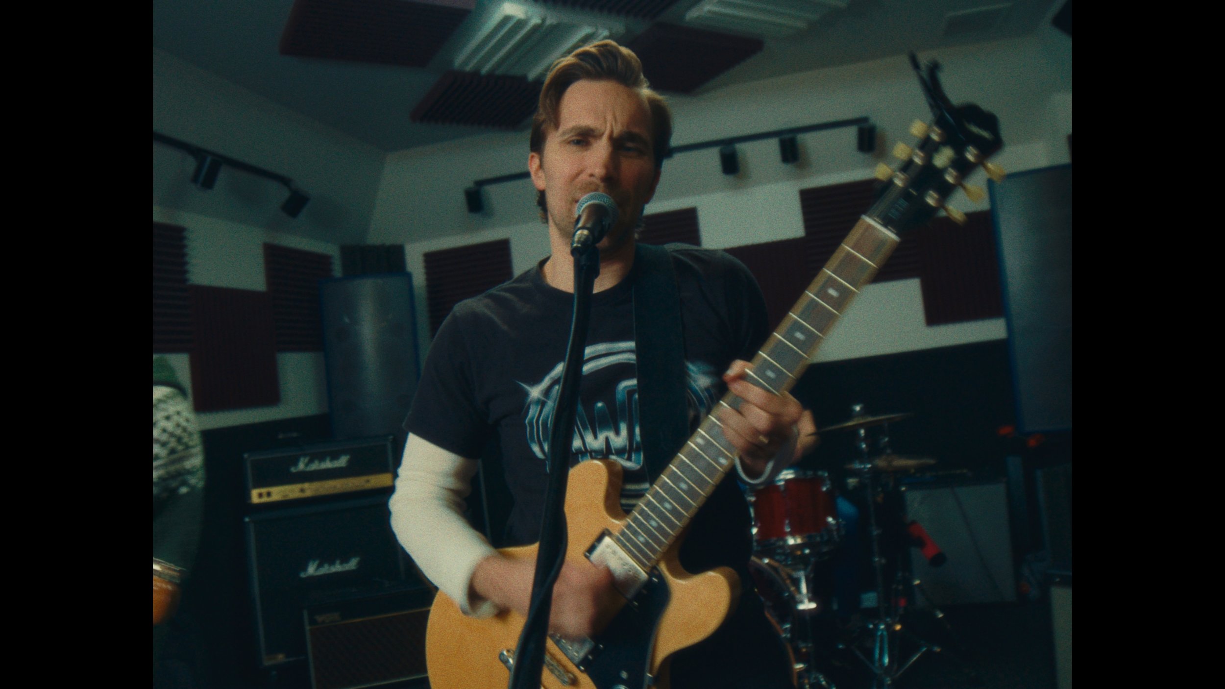 A man singing into a microphone and playing an electric guitar in a music rehearsal studio with amplifiers and drum set in the background.