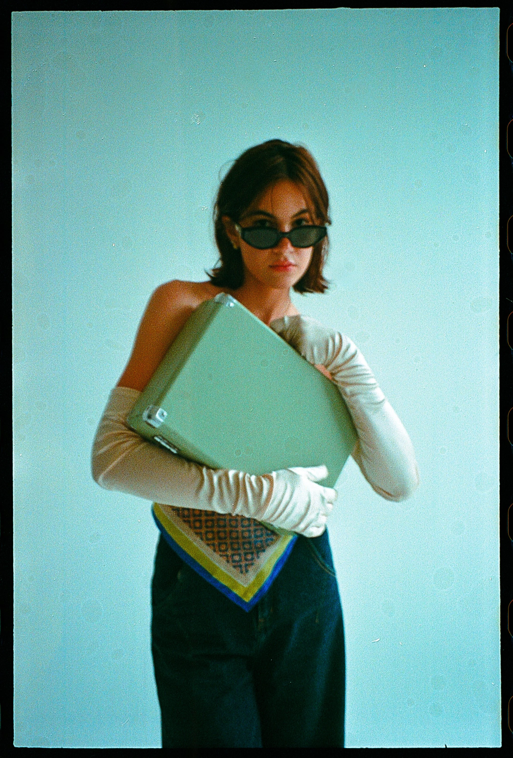 A woman with short brown hair wearing sunglasses, a white long-sleeve shirt, blue jeans, and a patterned scarf tied around her waist, holding a closed laptop against her chest, standing against a plain light blue background.