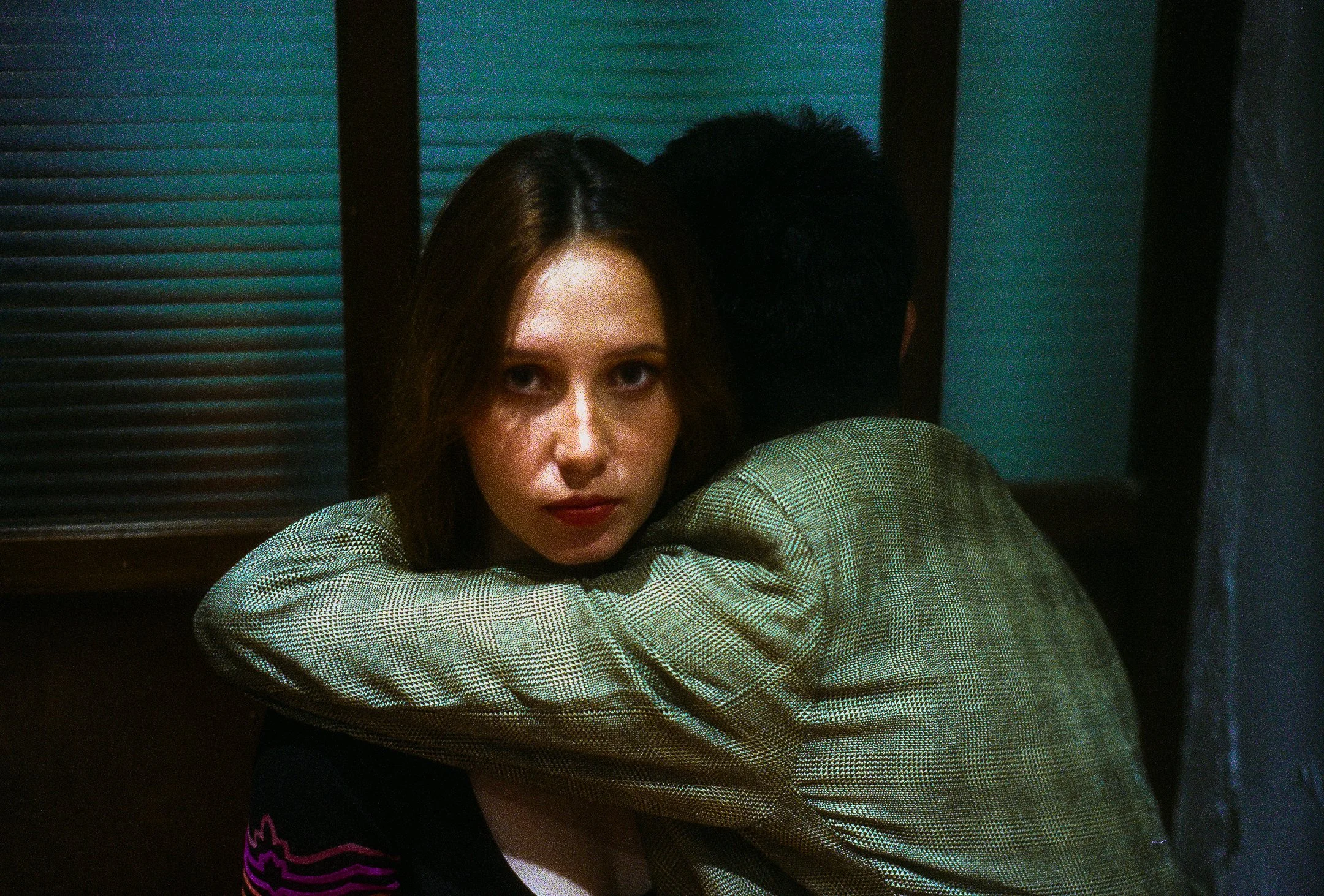 A woman with brown hair and red lipstick sitting with her arms around a man, facing the camera, in a dimly lit room with dark wooden walls and green window blinds.