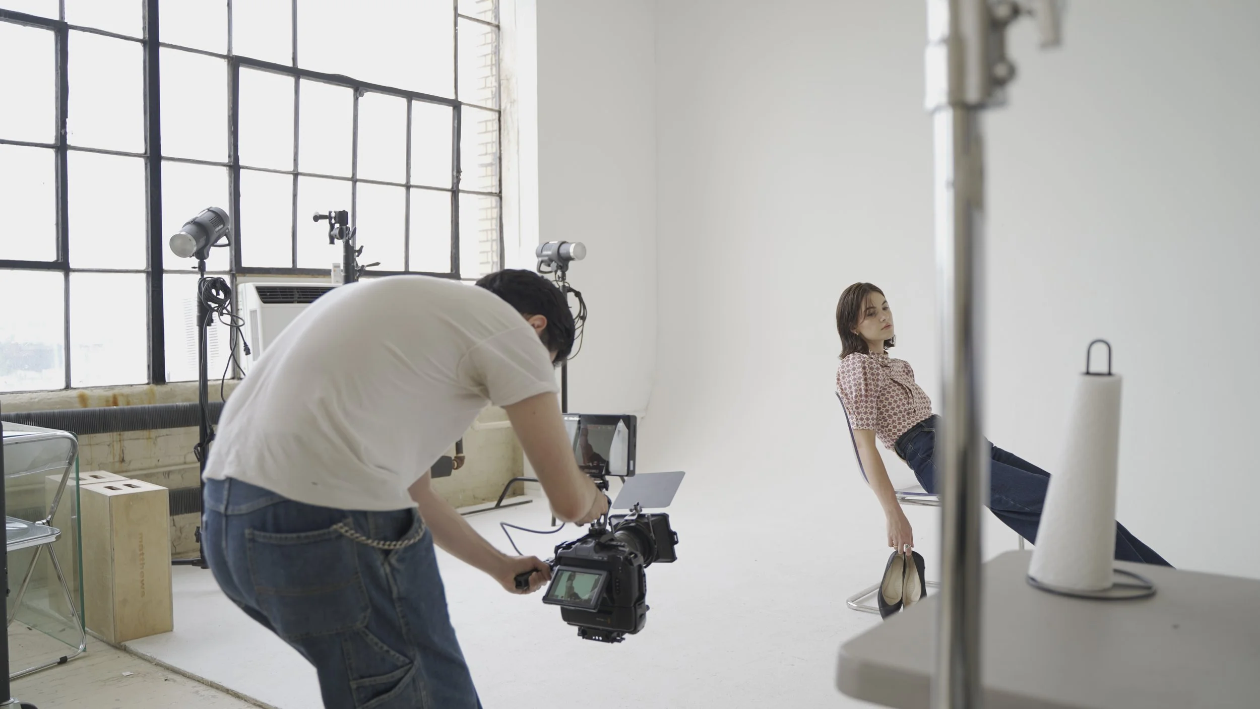 A model sitting on a metal chair in a minimalist photography studio with a white wall, holding a pair of shoes, while a photographer adjusts a camera on a tripod preparing for a photo shoot. Studio equipment like softbox lights and roll paper are visible.