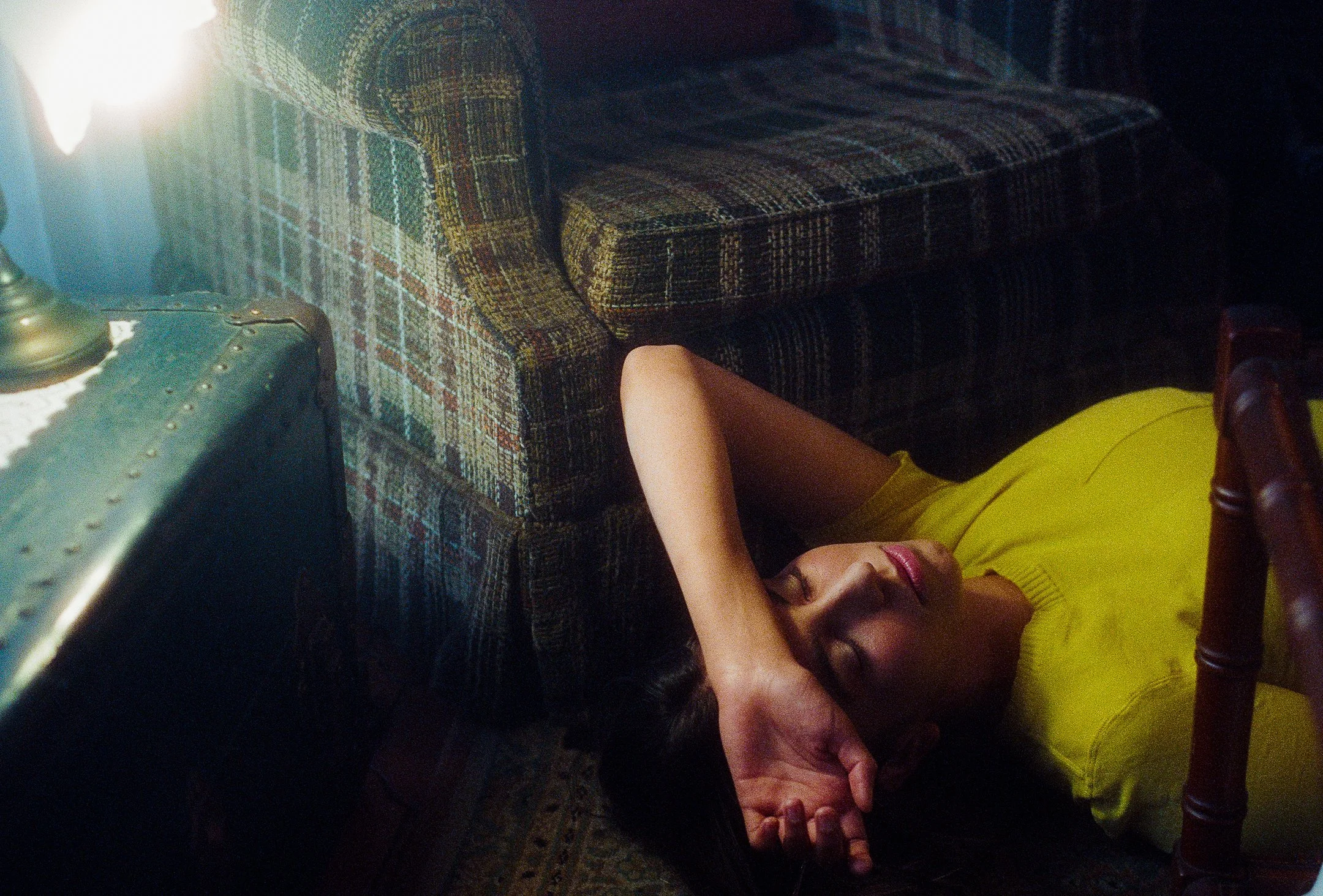 A woman wearing a yellow top lying on the floor with her eyes closed and arm over her forehead, next to a patterned sofa and a vintage lamp on a side table.