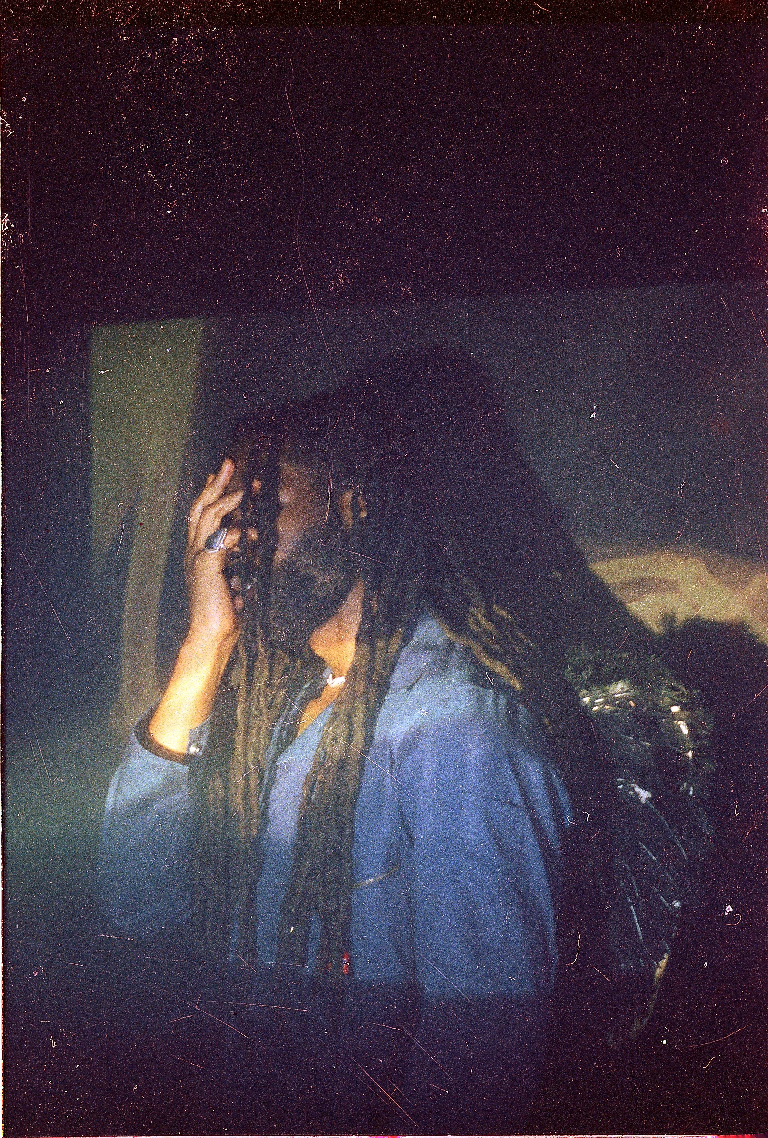 A person with long dreadlocks and dark clothing, touching their head, possibly adjusting hair or headphones, in a dimly lit room.