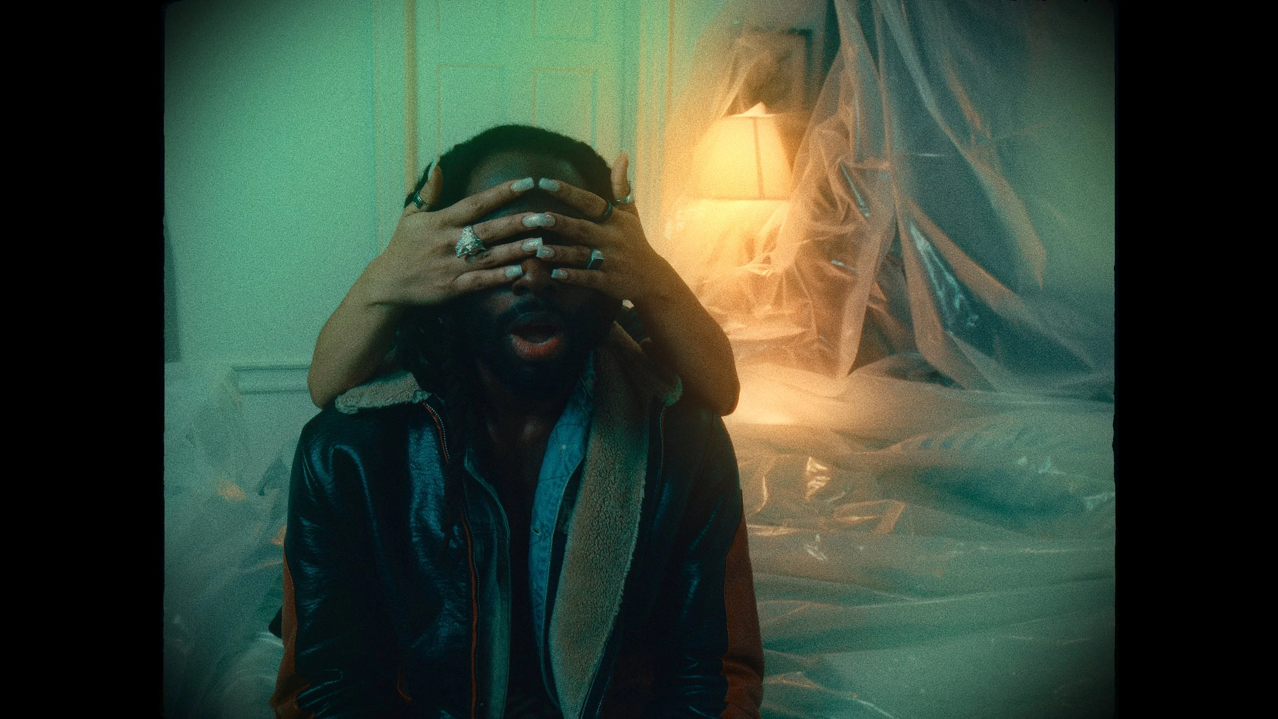 A person with glasses and a beard is sitting with their eyes covered by a second person who has hands adorned with rings resting on his face, in a dimly lit room with a lamp and plastic sheets in the background.