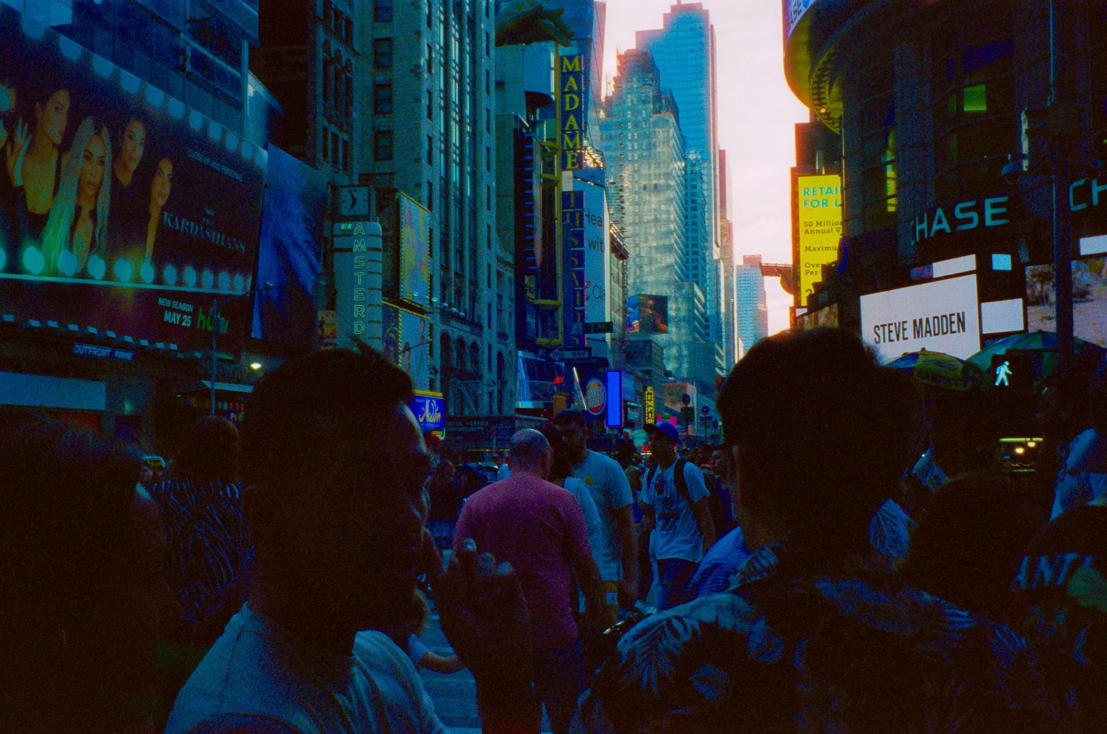 Crowded street scene in Times Square, New York City, during dusk with illuminated billboards and signs, including advertisements for 'The Kardashians' and 'Steve Madden', with people waiting at a pedestrian crosswalk.