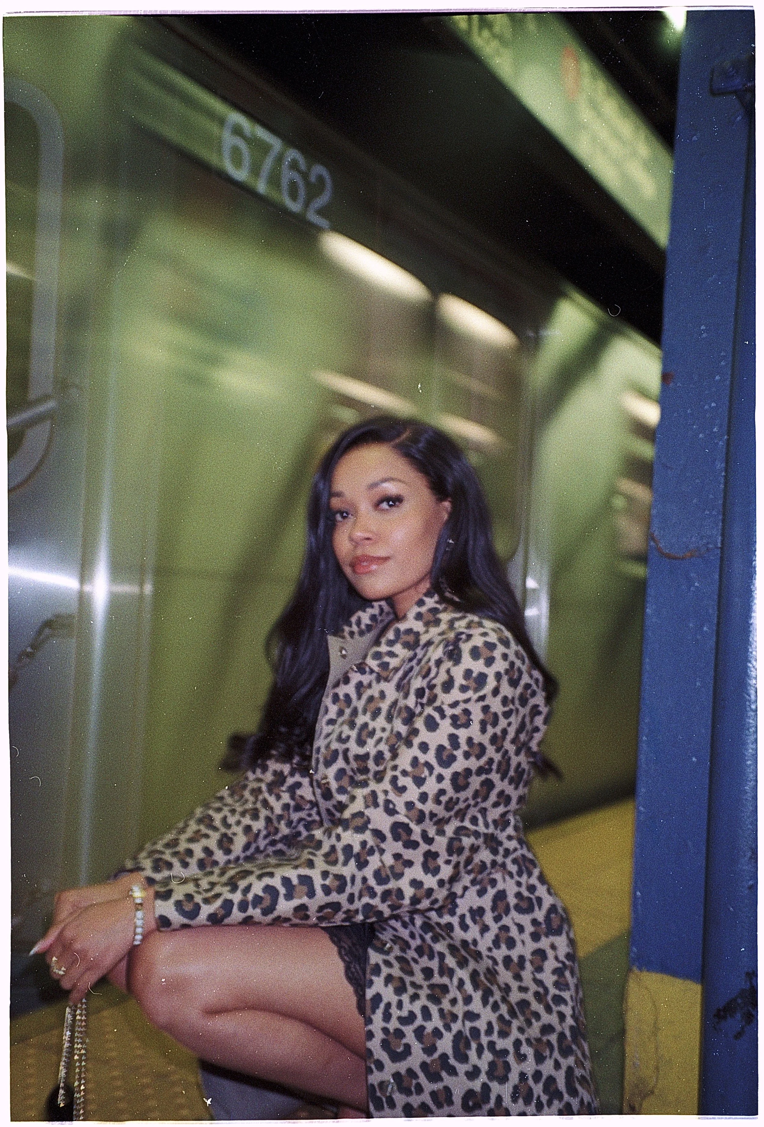 A woman with long dark hair sitting on the floor of an indoor area, wearing a leopard print coat and black lace leggings, with a neutral expression. She is accessorized with rings, bracelets, and earrings, and is looking slightly to the side. The background is a metallic green wall with a sign displaying the number "6762" and parts of an indoor transportation setting, possibly a subway or bus.