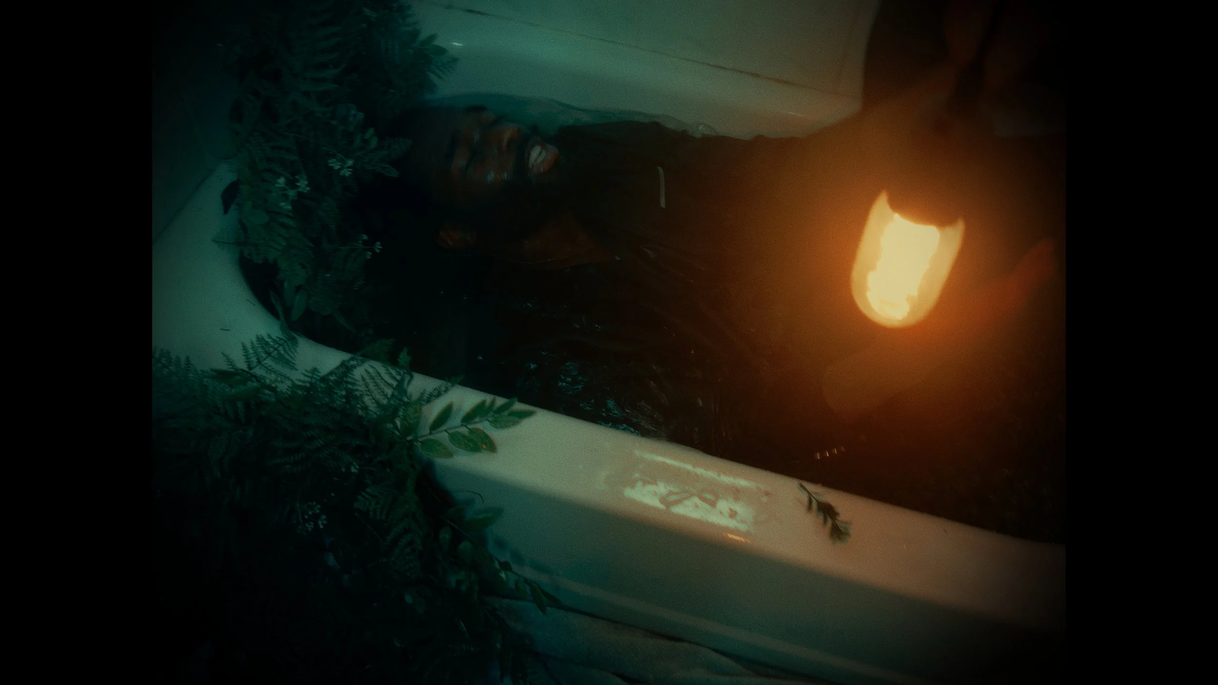 A person lying in a bathtub filled with water, surrounded by green plants, illuminated by a candle or fire.