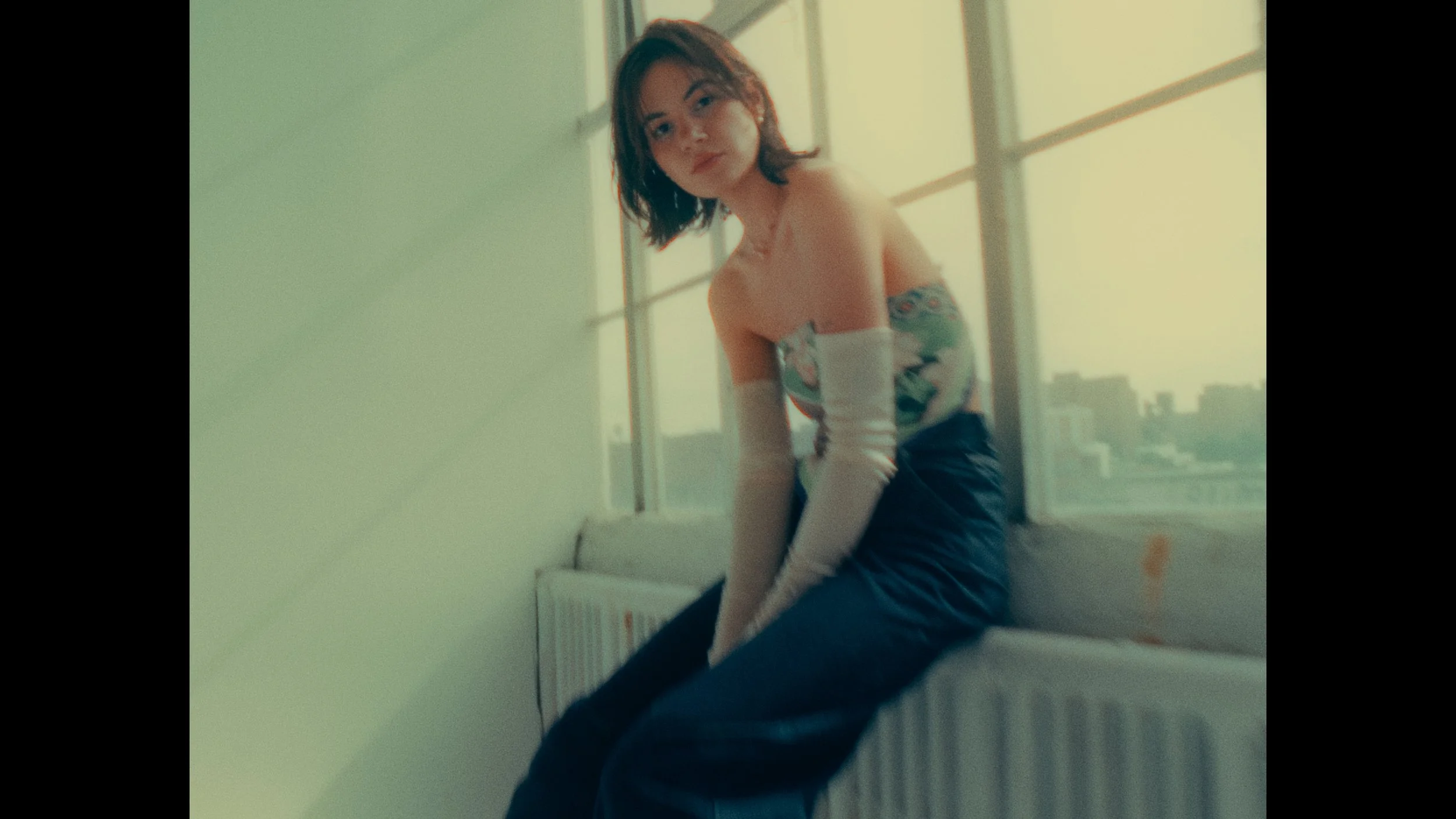 Young woman with shoulder-length hair sitting on a windowsill, wearing long light-colored gloves and an off-the-shoulder top with a floral design, posed indoors with cityscape visible through the window.