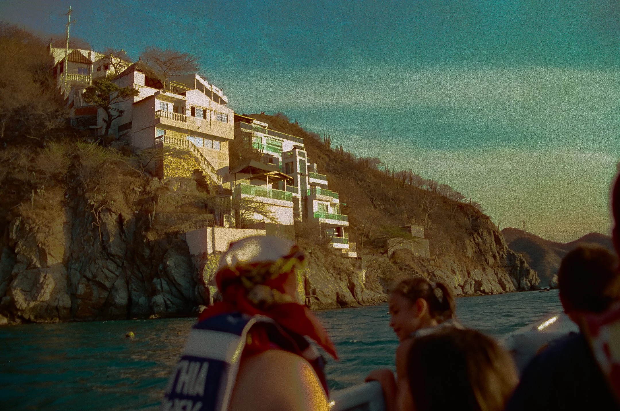 People on a boat near rocky coastline with hillside houses in the background, during sunset.