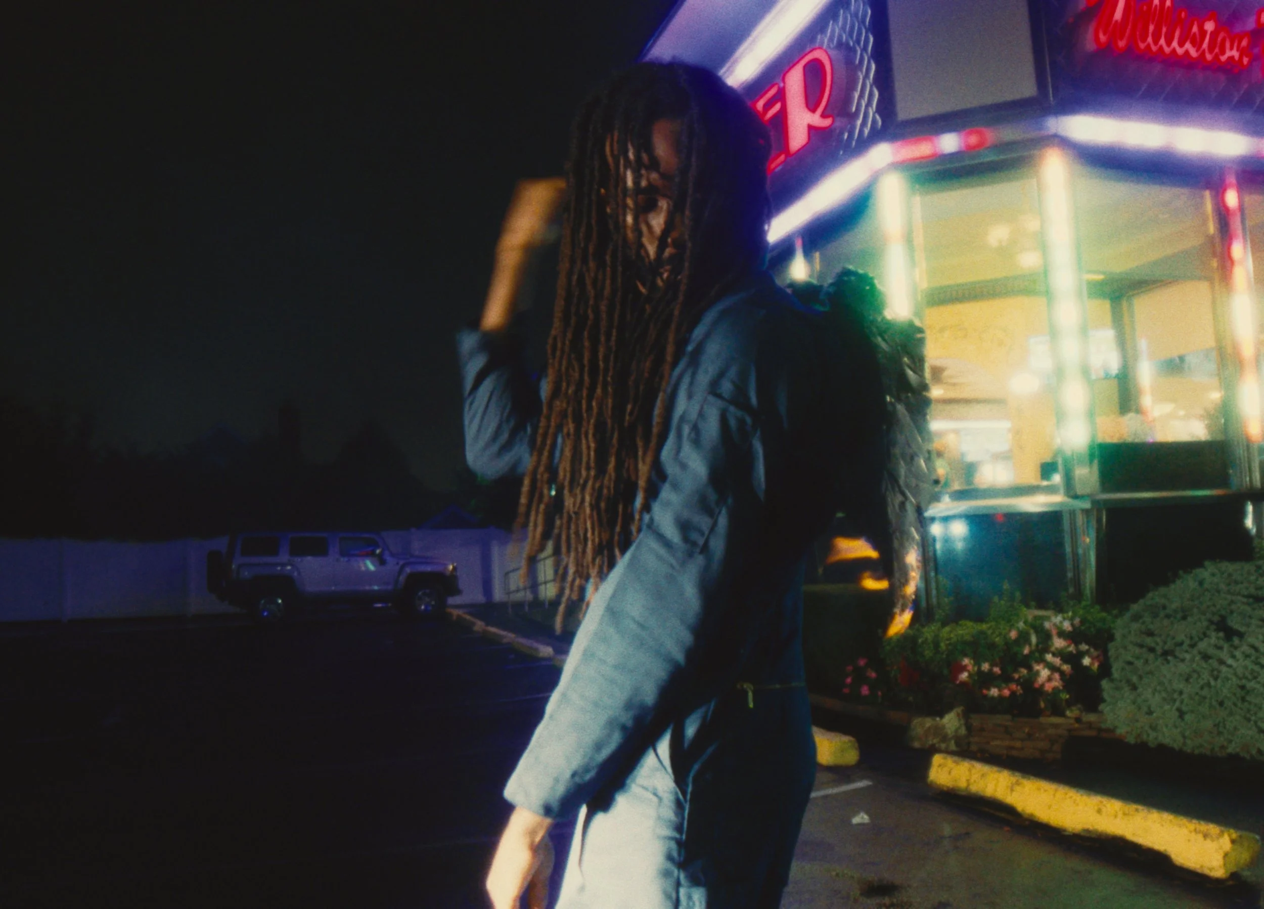 A person with long dreadlocks and a jean jacket stands at night in a parking lot with a brightly lit carnival or fair food stand in the background.