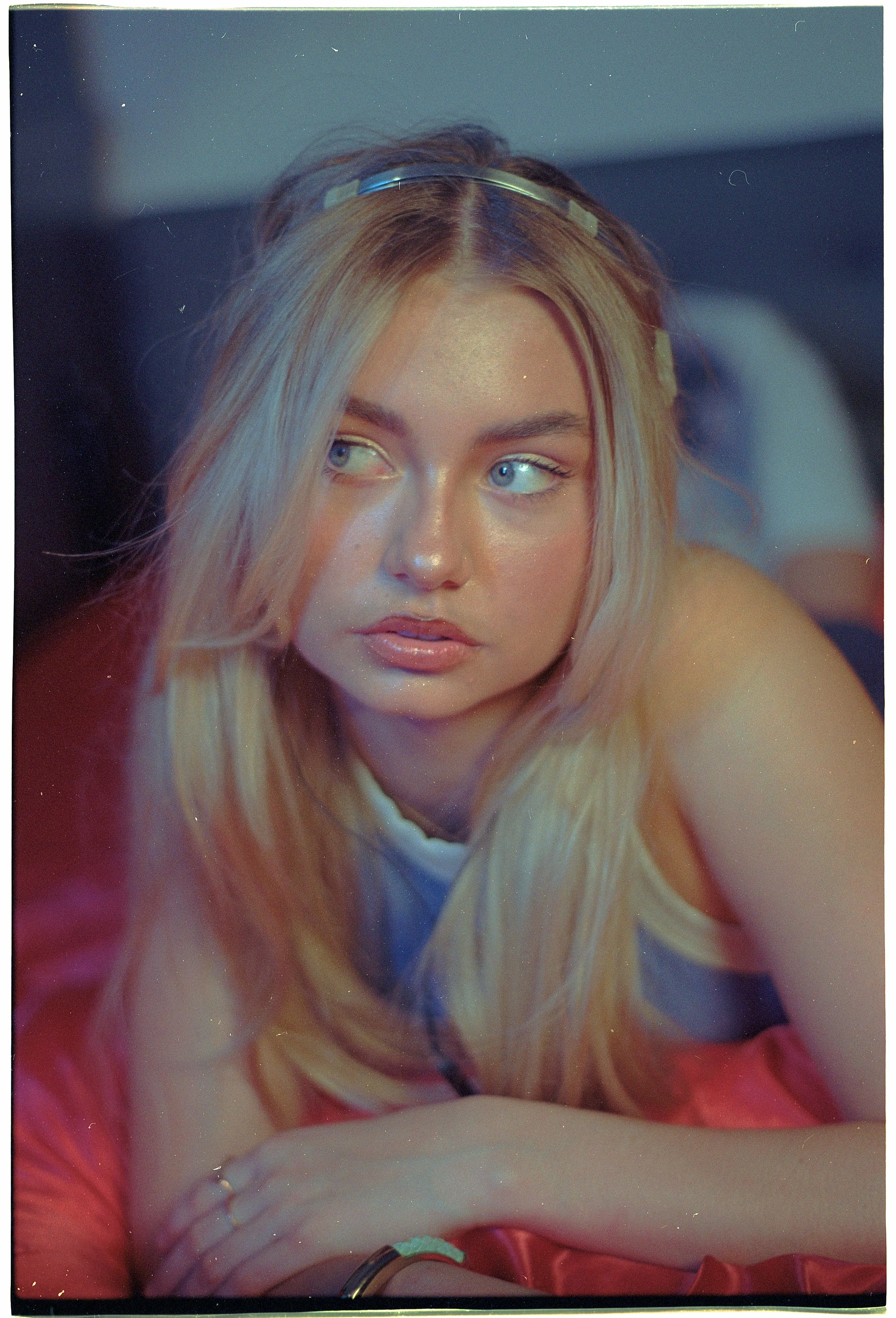 Close-up of a young woman with long blonde hair and blue eyes, wearing a metallic headband, resting her arm on a pink satin fabric, with a colorful soft-focus background. editorial photoshoot for a brand