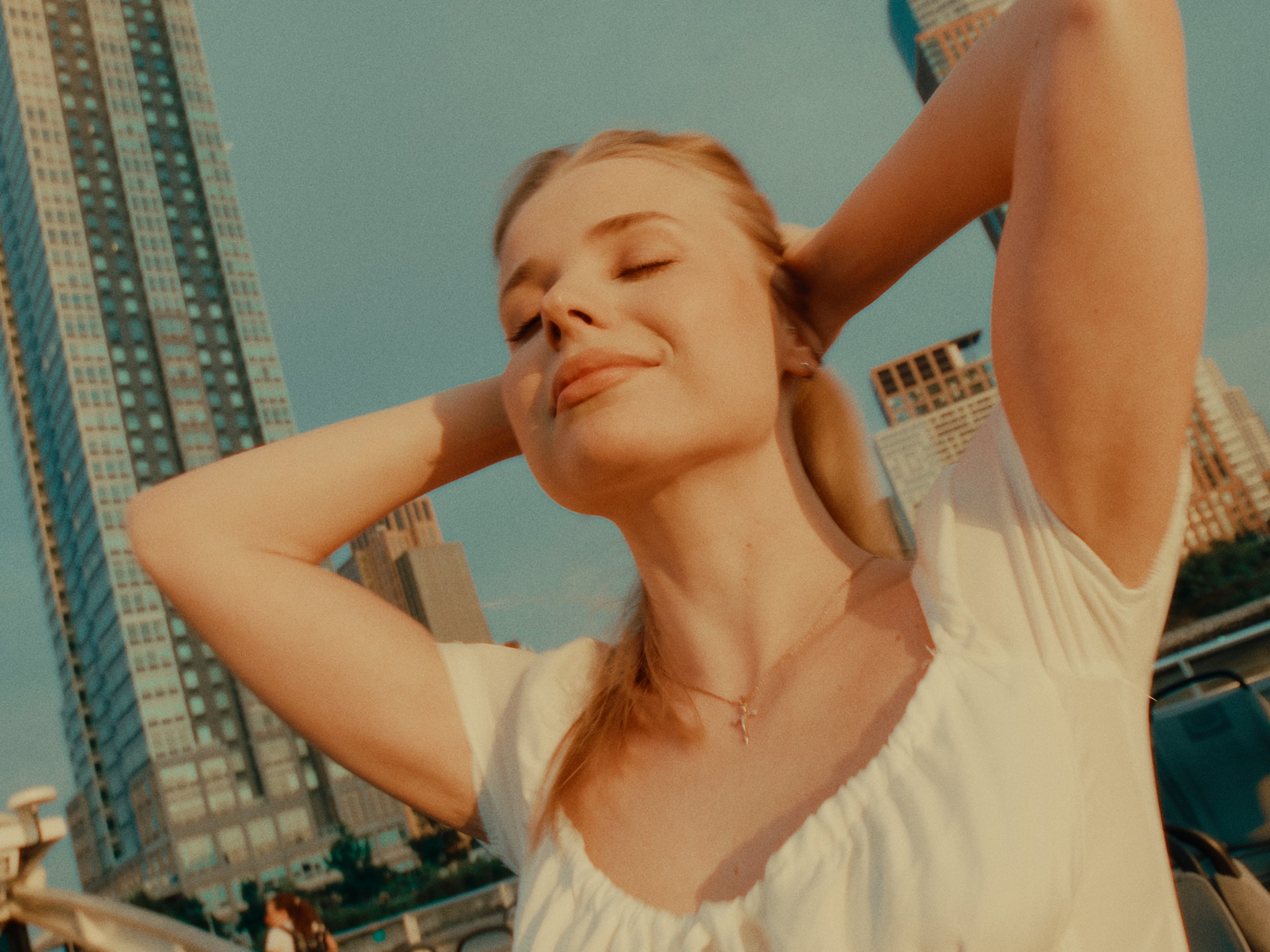 Young woman with blonde hair relaxing outdoors with skyscrapers in the background, eyes closed, smiling, wearing a white top and a necklace.