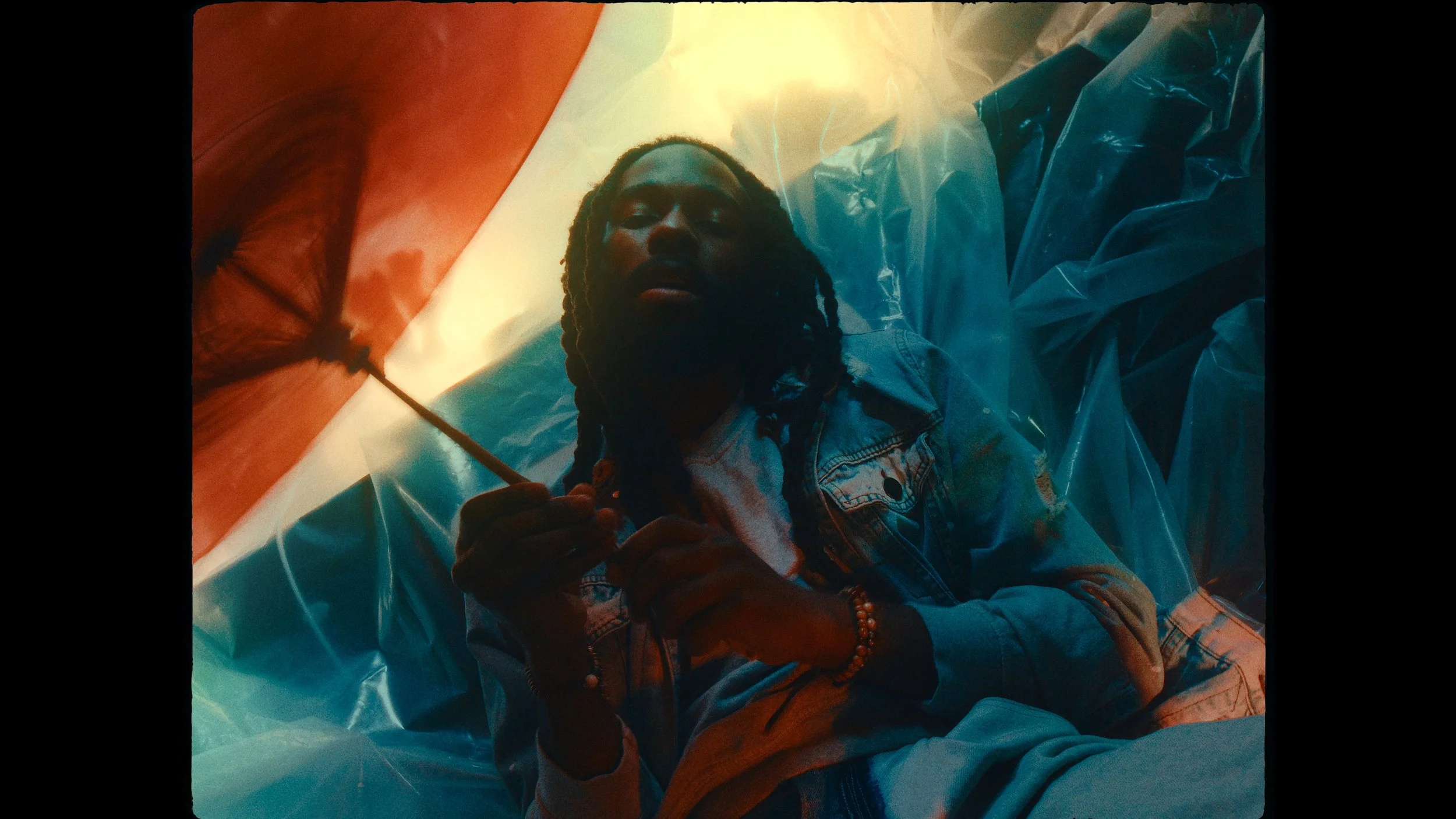 A person with long dreadlocks and a denim jacket lying down on a surface covered with plastic, holding a lit cigarette, with a colorful abstract background and light effects.