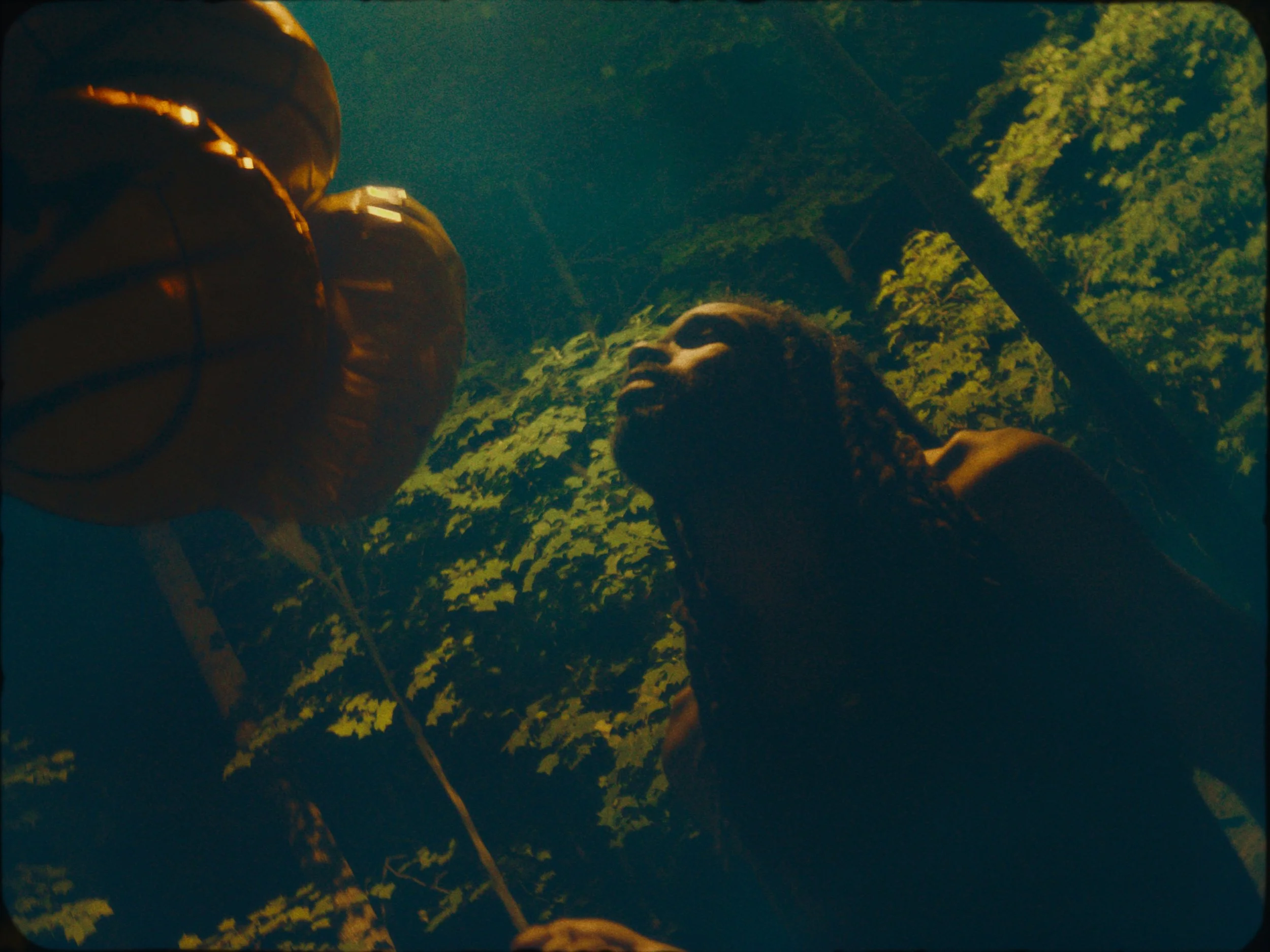 A person with long hair is standing outdoors among trees, looking at two orange balloons tied together with a string.