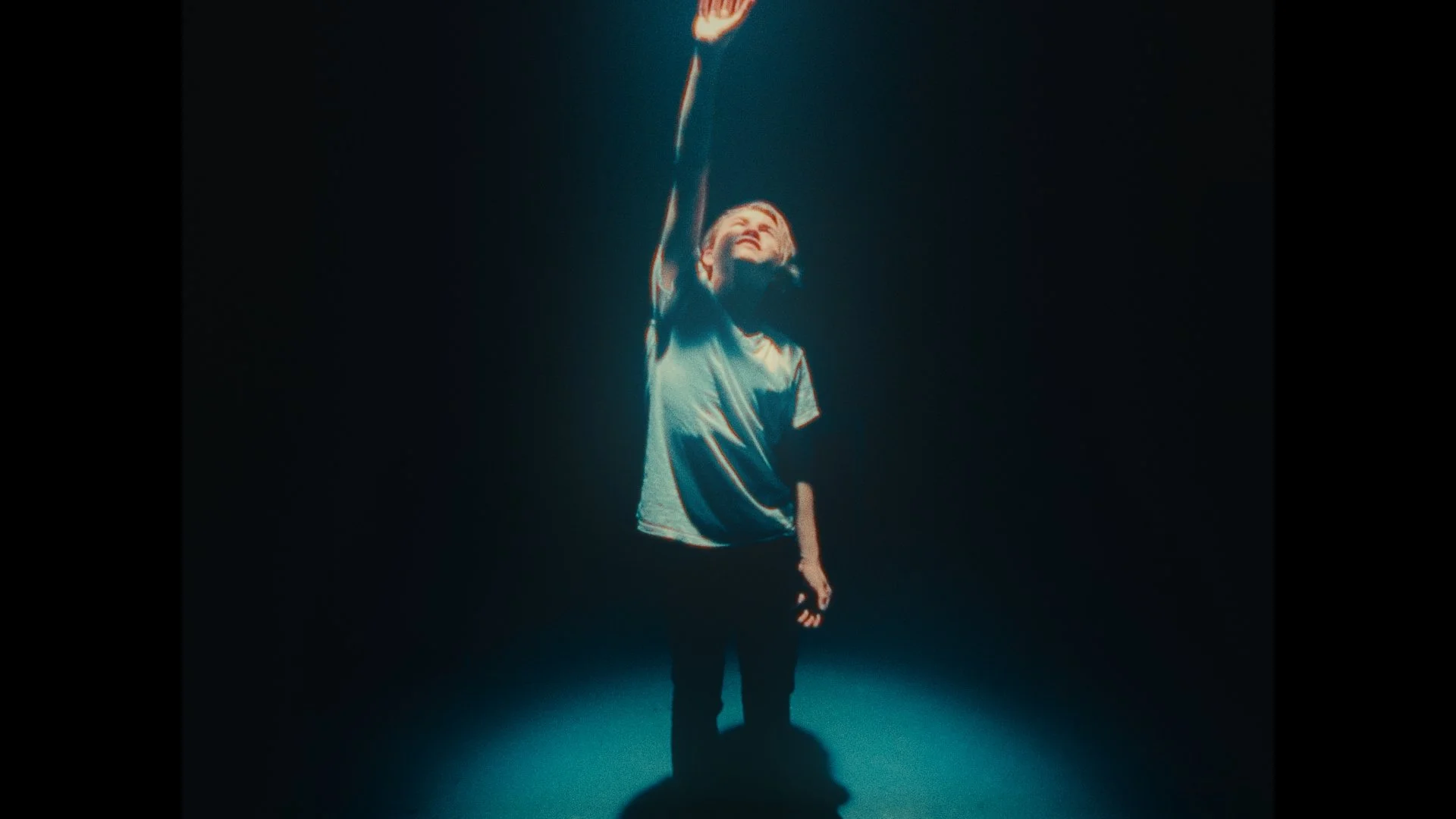 A young person with short hair, wearing a shiny silver shirt, stands in a dark space with a spotlight illuminating them from below, looking upward with a joyful expression and one arm raised.